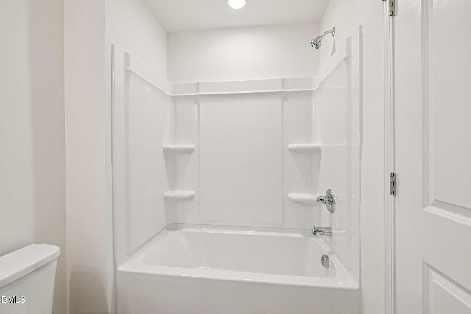 White bathroom with a built-in bathtub and shower combination, featuring four built-in shelves. The walls and ceiling are painted white. There is a closed white door to the right and a partial view of a toilet to the left.