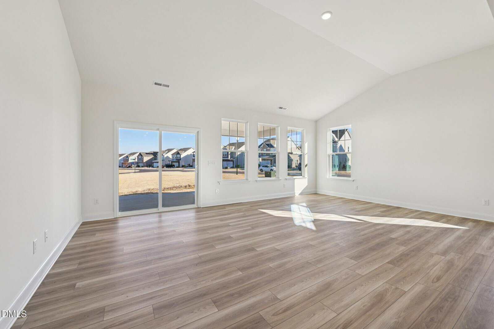 Bright, empty room with light wood flooring, white walls, large windows, and glass doors opening to a patio. Sunlight streams in, and houses are visible outside. The ceiling is vaulted with recessed lighting.
