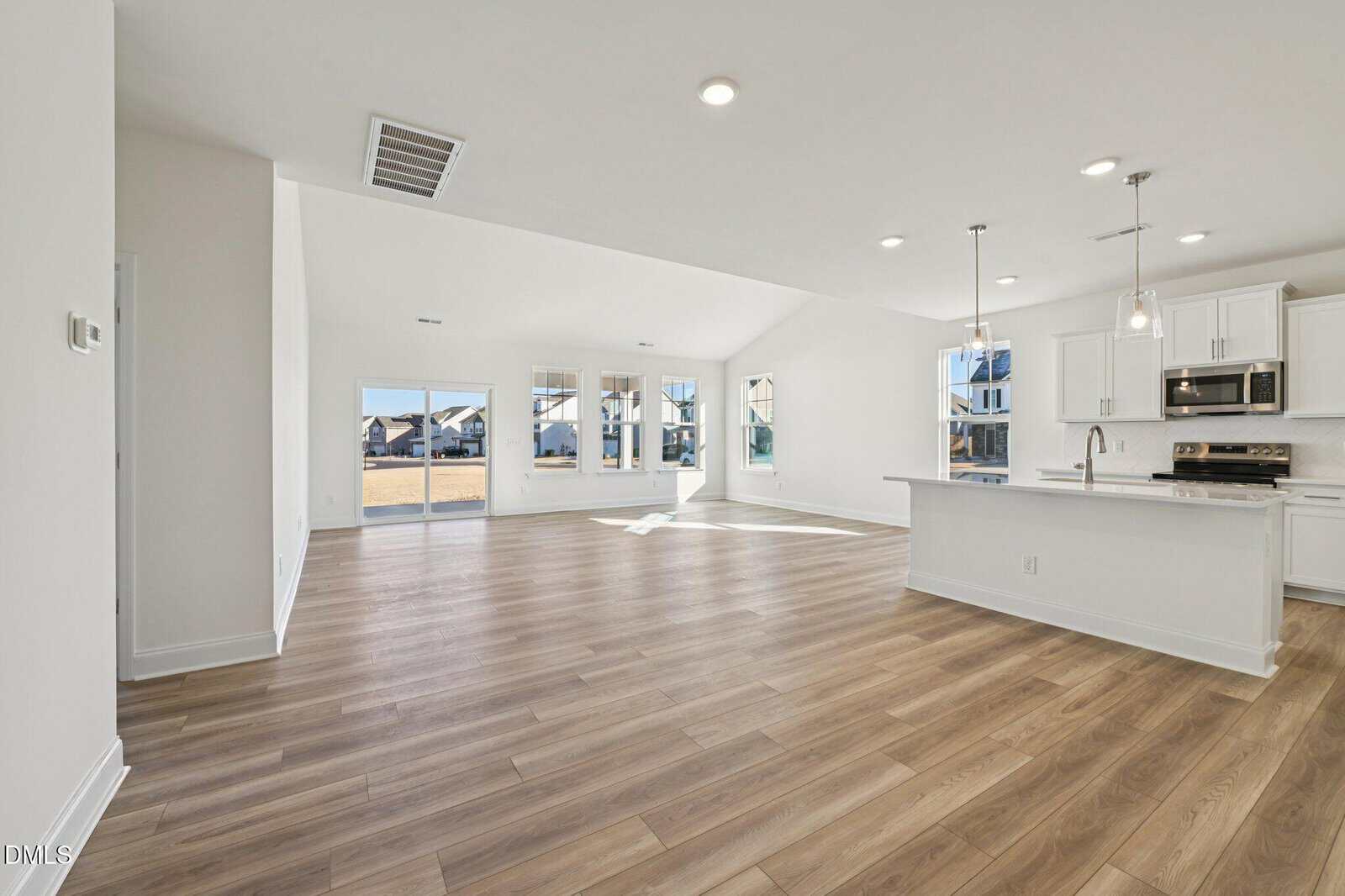 Spacious open-concept living area with large windows, light wood flooring, and a modern white kitchen featuring an island, stainless steel appliances, and pendant lighting. Bright and airy with ample natural light.