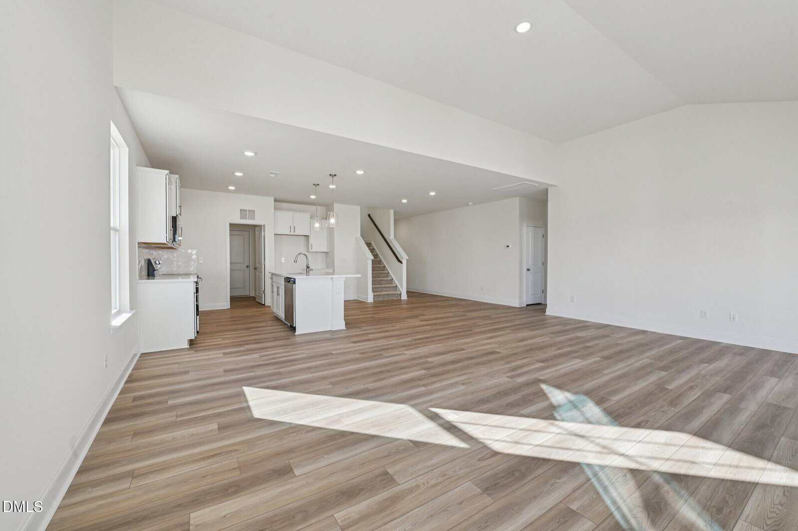 Open-concept living area with light wood flooring, white walls, and recessed lighting. Modern kitchen with island, white cabinets, and stainless steel appliances, plus a staircase leading to an upper level. Large window casts shadows on floor.