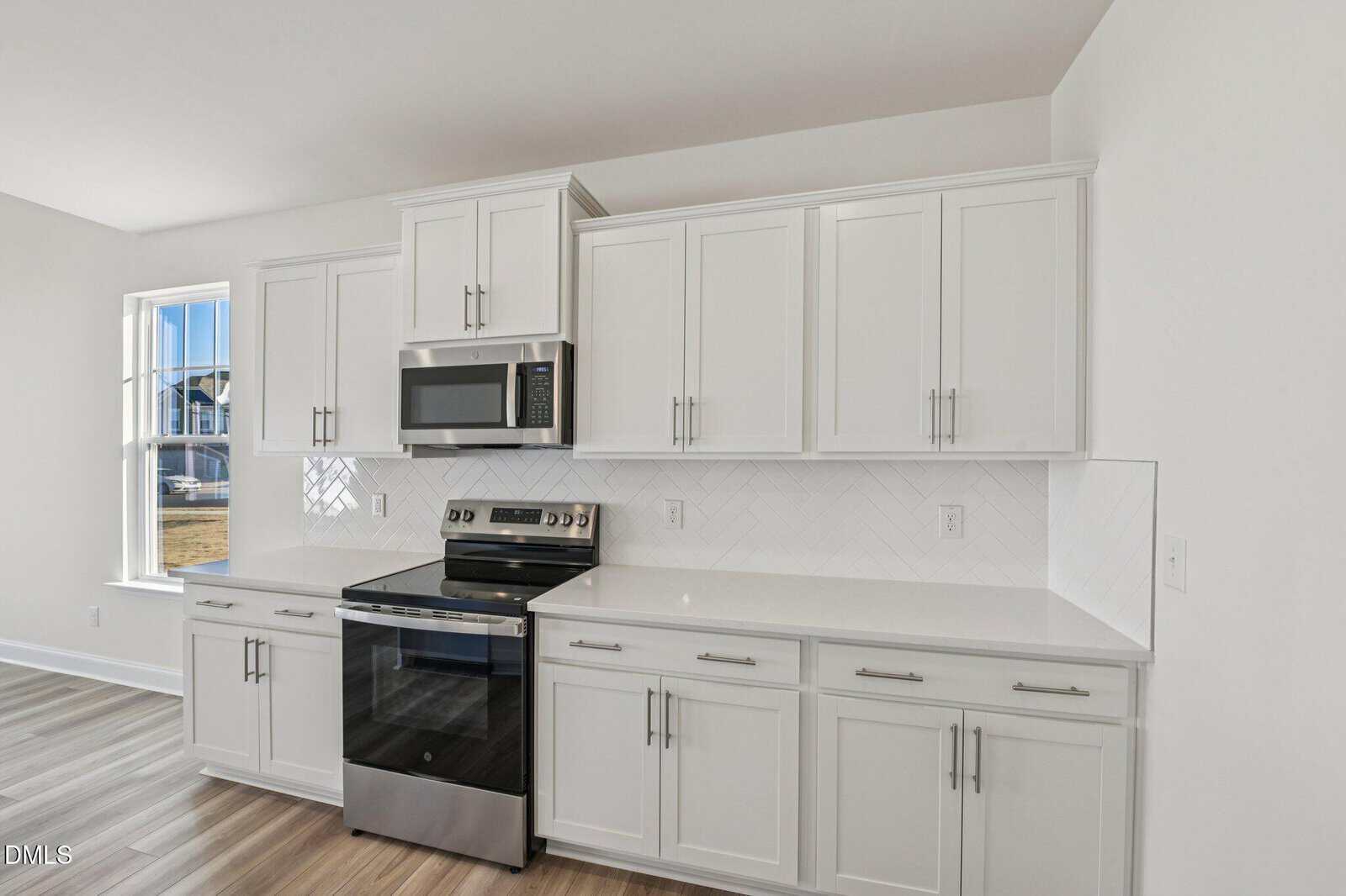Modern kitchen with white cabinets, stainless steel oven and microwave, white countertops, herringbone tile backsplash, and light wood flooring. A window is on the left, letting in natural light.