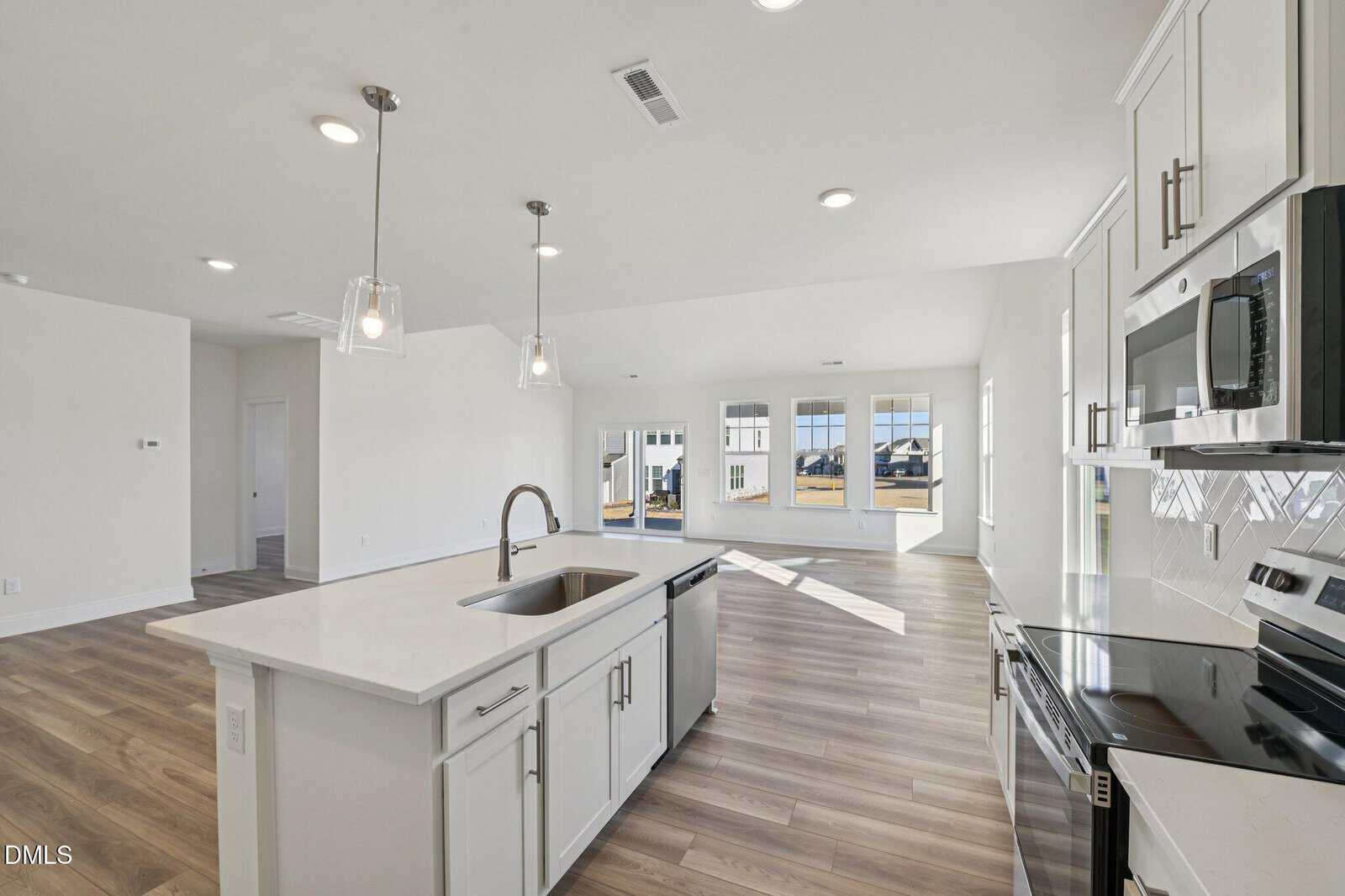 Modern open-concept kitchen and living area with white cabinetry, stainless steel appliances, pendant lights over an island, light wood flooring, and large windows letting in natural light.