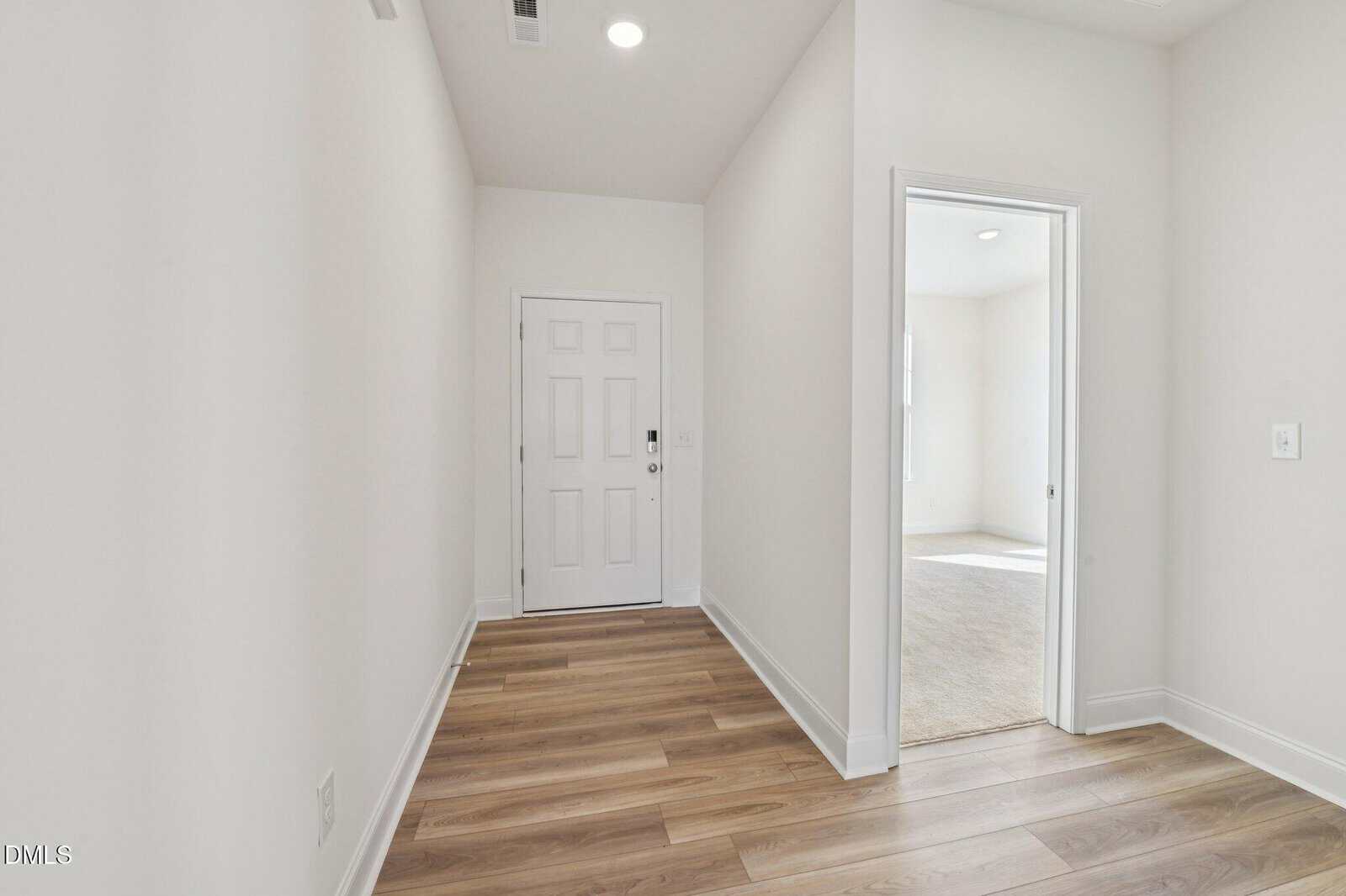 A bright, empty hallway with light wood flooring, white walls, and a closed white front door. An open doorway on the right leads to a carpeted room filled with natural light.