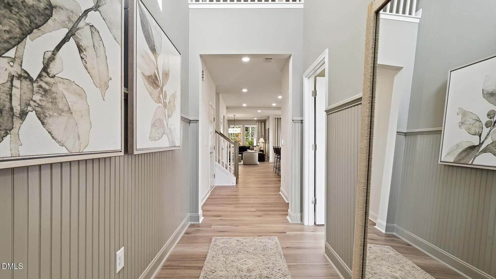 A hallway with light wood flooring, gray paneled walls, botanical artwork, a large wall mirror, and a small rug, leading to a bright living area with recessed lighting and visible furniture in the distance.