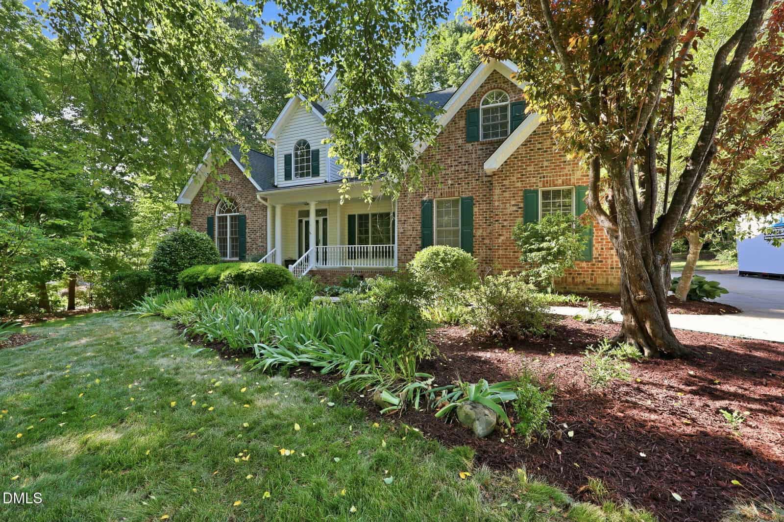 A two-story brick house with white trim, green shutters, a covered front porch, and manicured landscaping with shrubs, trees, and a flower bed in a lush, green yard.