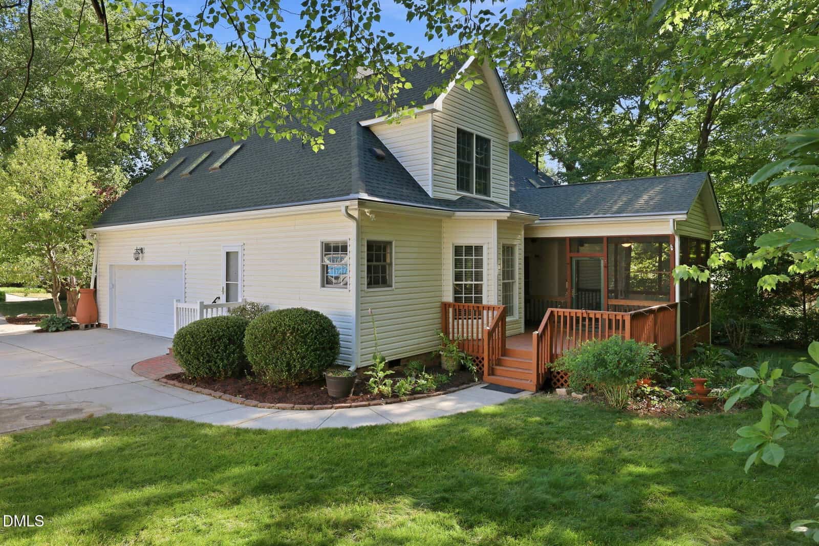 A two-story light yellow house with a dark roof, attached two-car garage, screened porch, and wooden steps, surrounded by trees, green grass, and landscaped bushes on a sunny day.