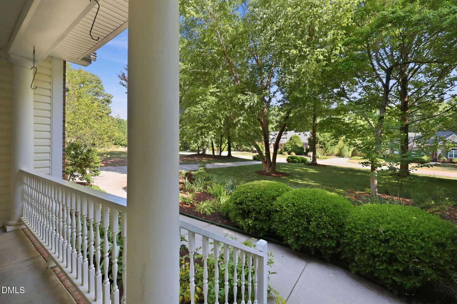 View from a front porch with white railing and columns, looking out onto a green yard with manicured bushes, trees, and a driveway. Houses are visible in the background under a clear blue sky.