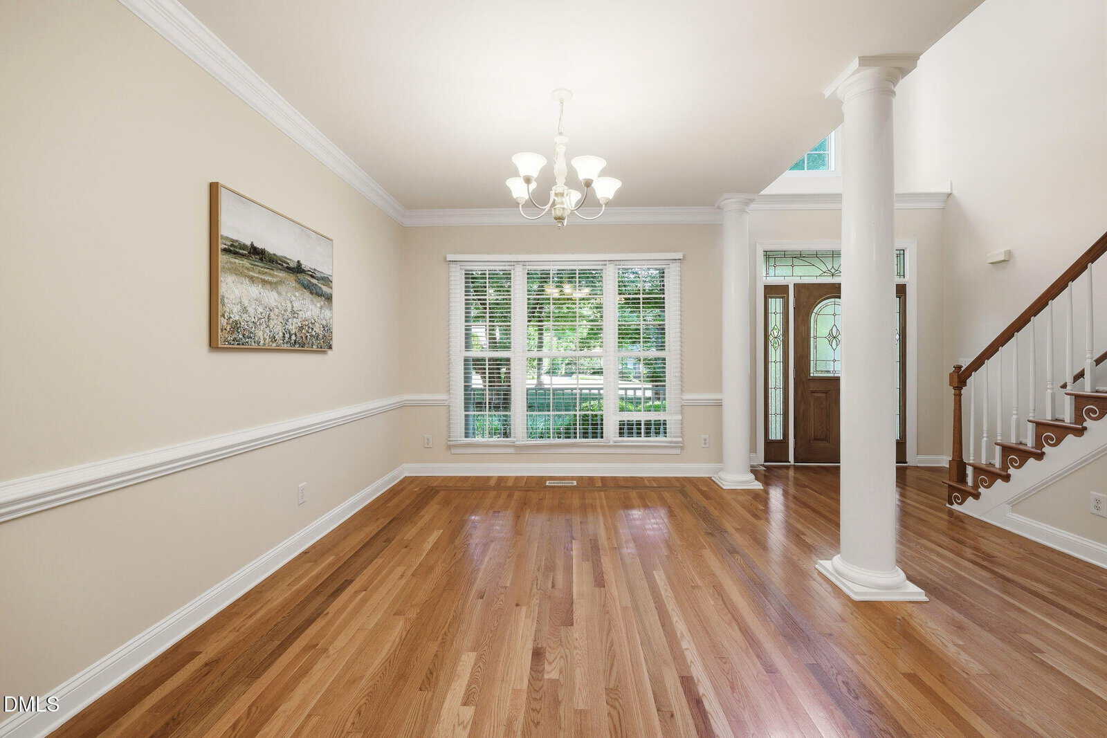 Bright, empty room with hardwood floors, large window with blinds, chandelier, white walls, front door, staircase with wooden steps, and a column near the entrance. A landscape painting hangs on the left wall.