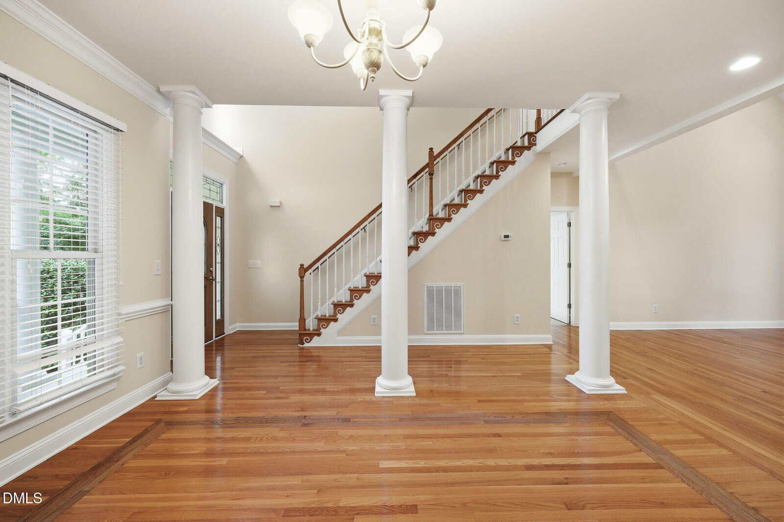 Bright, open living space with hardwood floors, white columns, a staircase with wooden railing, large windows with blinds, and a modern chandelier. Cream walls and baseboards create a clean, airy feel.