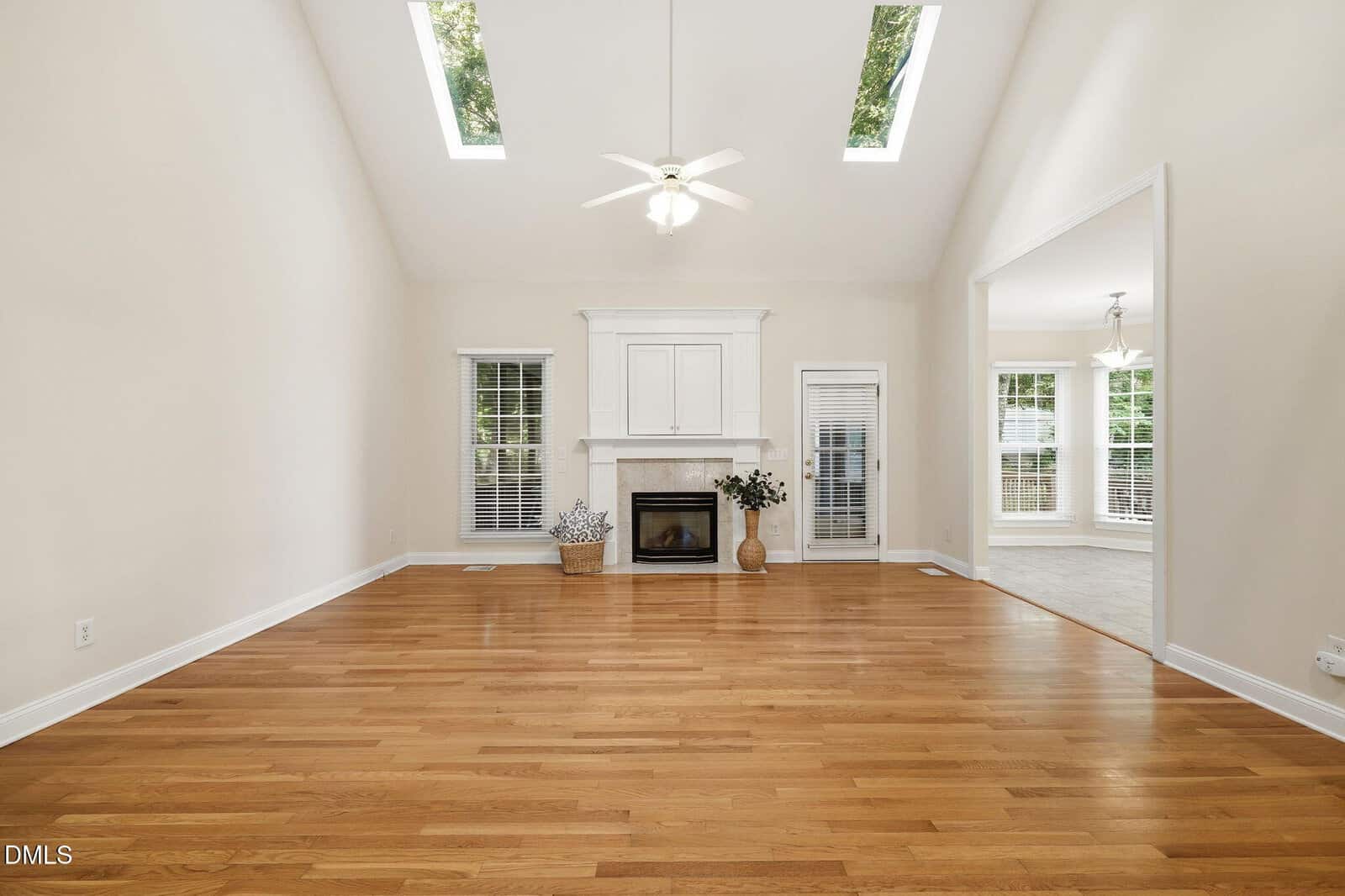 Spacious living room with light wood floors, a white fireplace, high ceiling, skylights, and large windows. An adjacent room with tiled floors is visible through a wide doorway.
