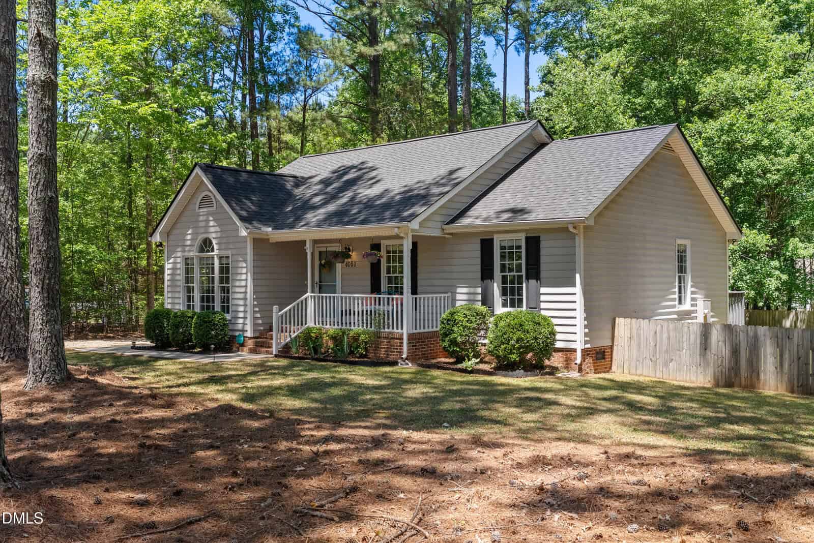 Single-story light gray house with white trim, a front porch, black shutters, and a gabled roof, surrounded by trees and a grassy yard with a wooden fence to the right.
