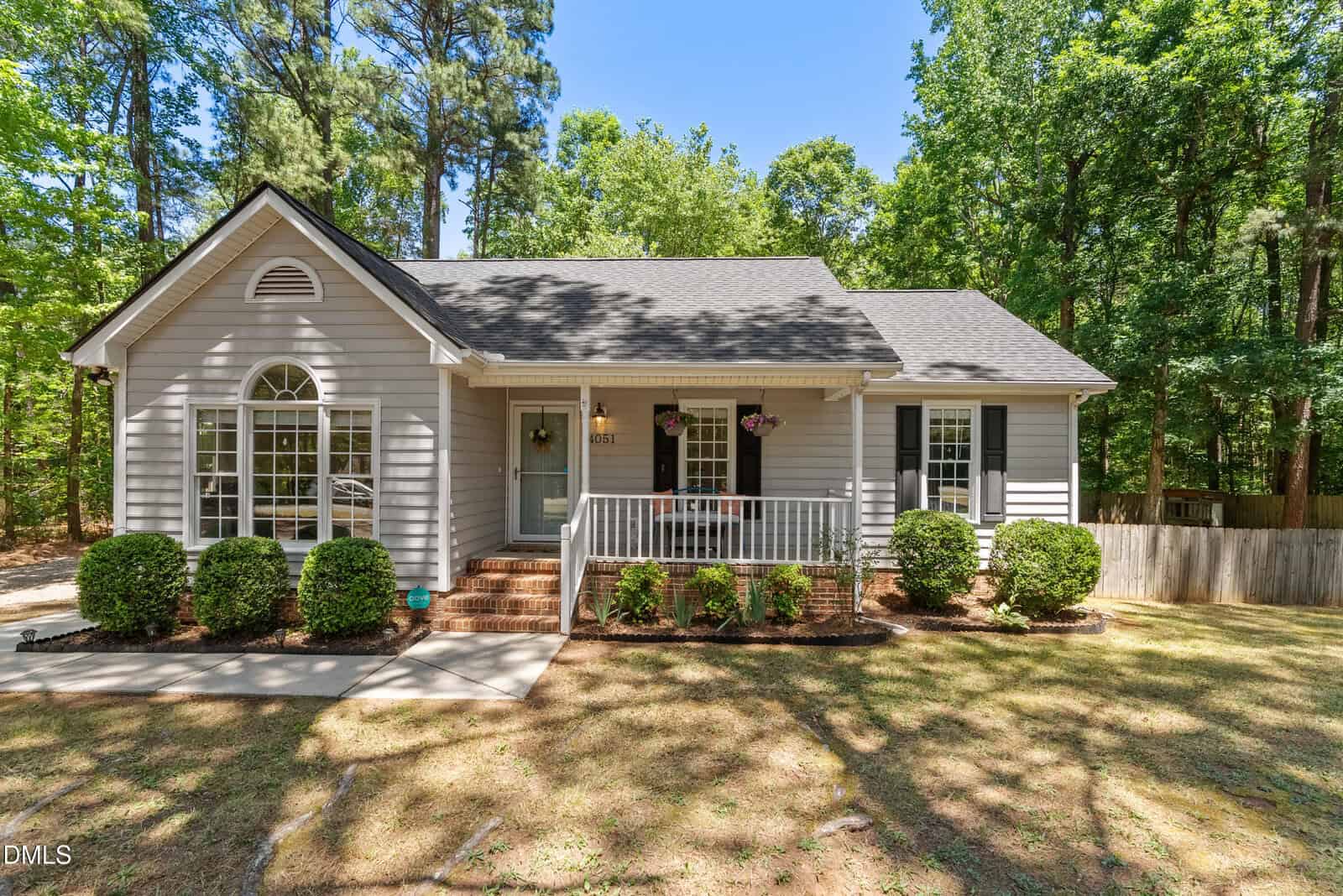 A single-story, light gray house with white trim sits at 4051 Old Franklinton Road, featuring a front porch, hanging flower baskets, arched window, black shutters, manicured shrubs, and tall trees framed by a wooden fence in Franklinton.