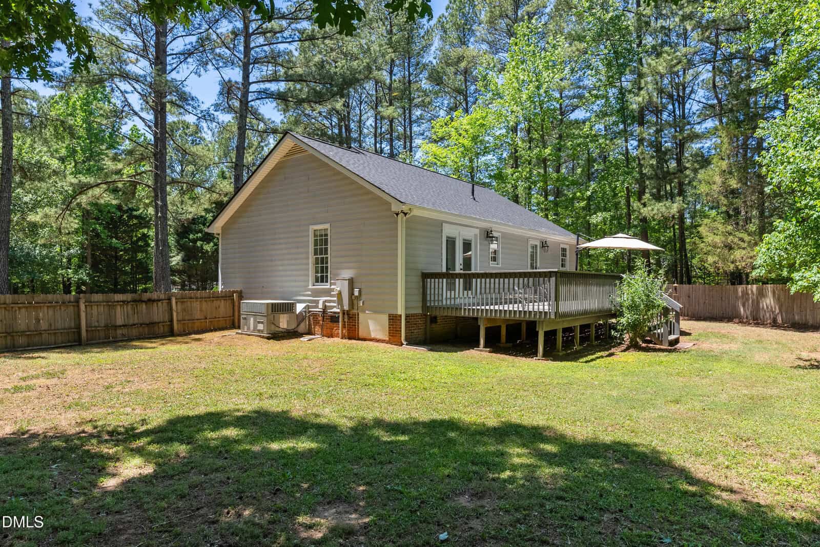 A single-story light gray house with a fenced backyard, wooden deck, and outdoor umbrella, surrounded by green grass, trees, and shrubs on a sunny day.