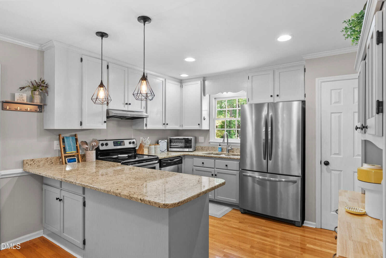 Modern kitchen with white cabinets, stainless steel appliances, granite countertops, pendant lights over an island, wooden floors, a window above the sink, and light gray walls.