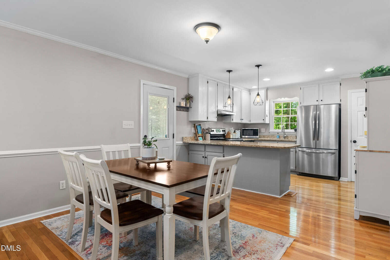 Modern kitchen and dining area with a wooden table, four white chairs, light gray cabinets, stainless steel appliances, pendant lights, and a colorful rug on polished wood flooring.
