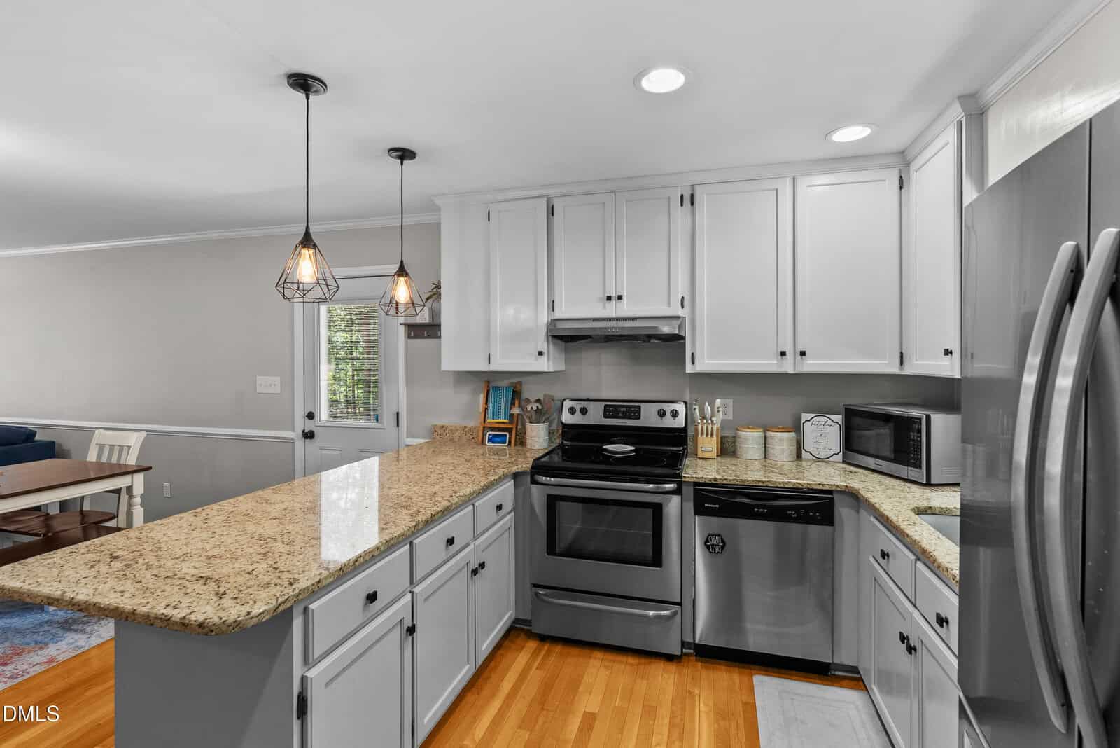 Modern kitchen with stainless steel appliances, granite countertops, white cabinets, pendant lighting, and hardwood floors. Dining area visible in the background.