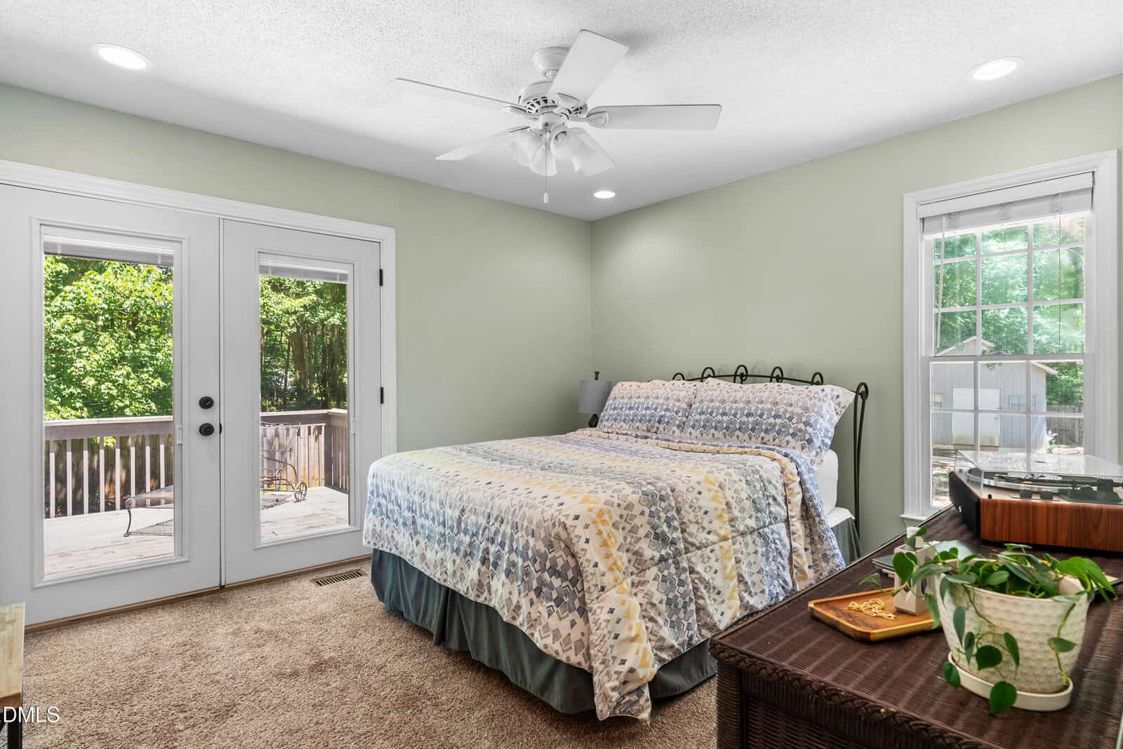 A cozy bedroom with light green walls, a bed with a patterned quilt, a window with outdoor views, double glass doors leading to a deck, carpeted floor, a dresser, and a potted plant.