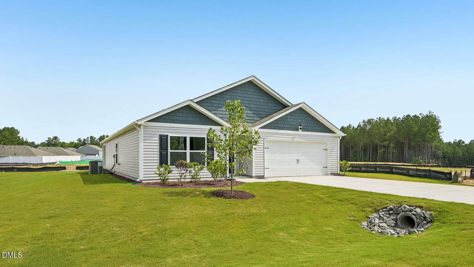 Single-story suburban house with gray siding, white trim, a two-car garage, small trees, and a manicured lawn. The driveway slopes toward a drain pipe, and there are wooded areas in the background under a clear sky.