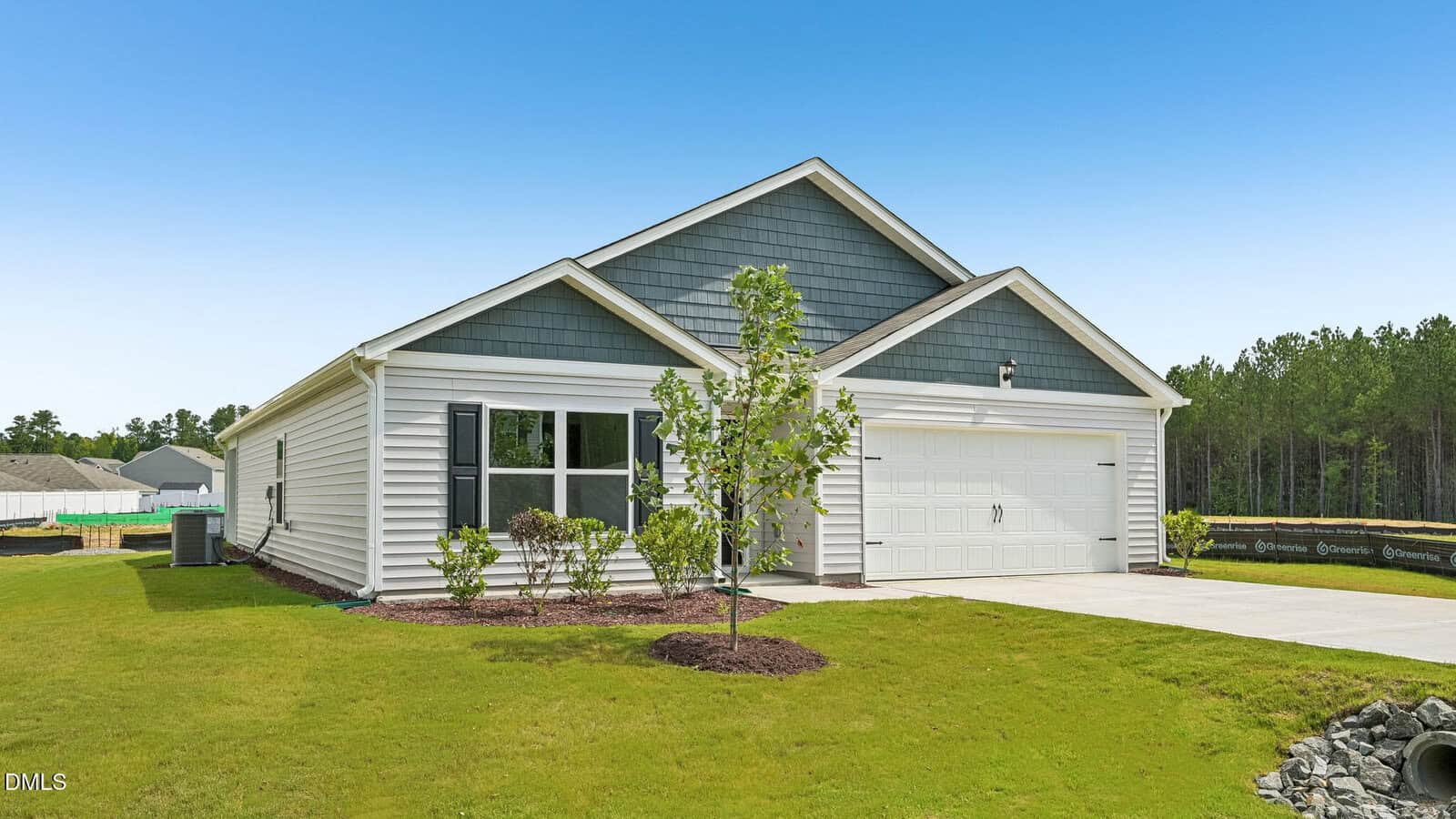 Single-story house with gray siding and white trim, two-car garage, front lawn, small tree, shrubs, and a backdrop of trees under a clear blue sky.