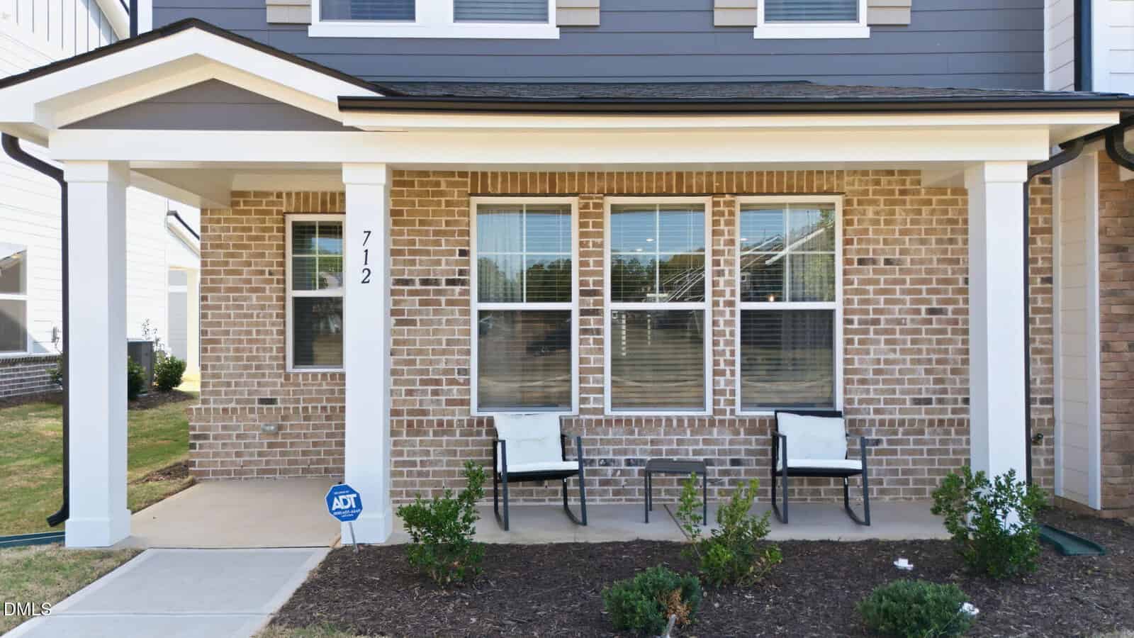 A brick house with a covered front porch featuring two white chairs, a small table, and an ADT security sign. Three large windows face the porch, and shrubs are planted along the front.