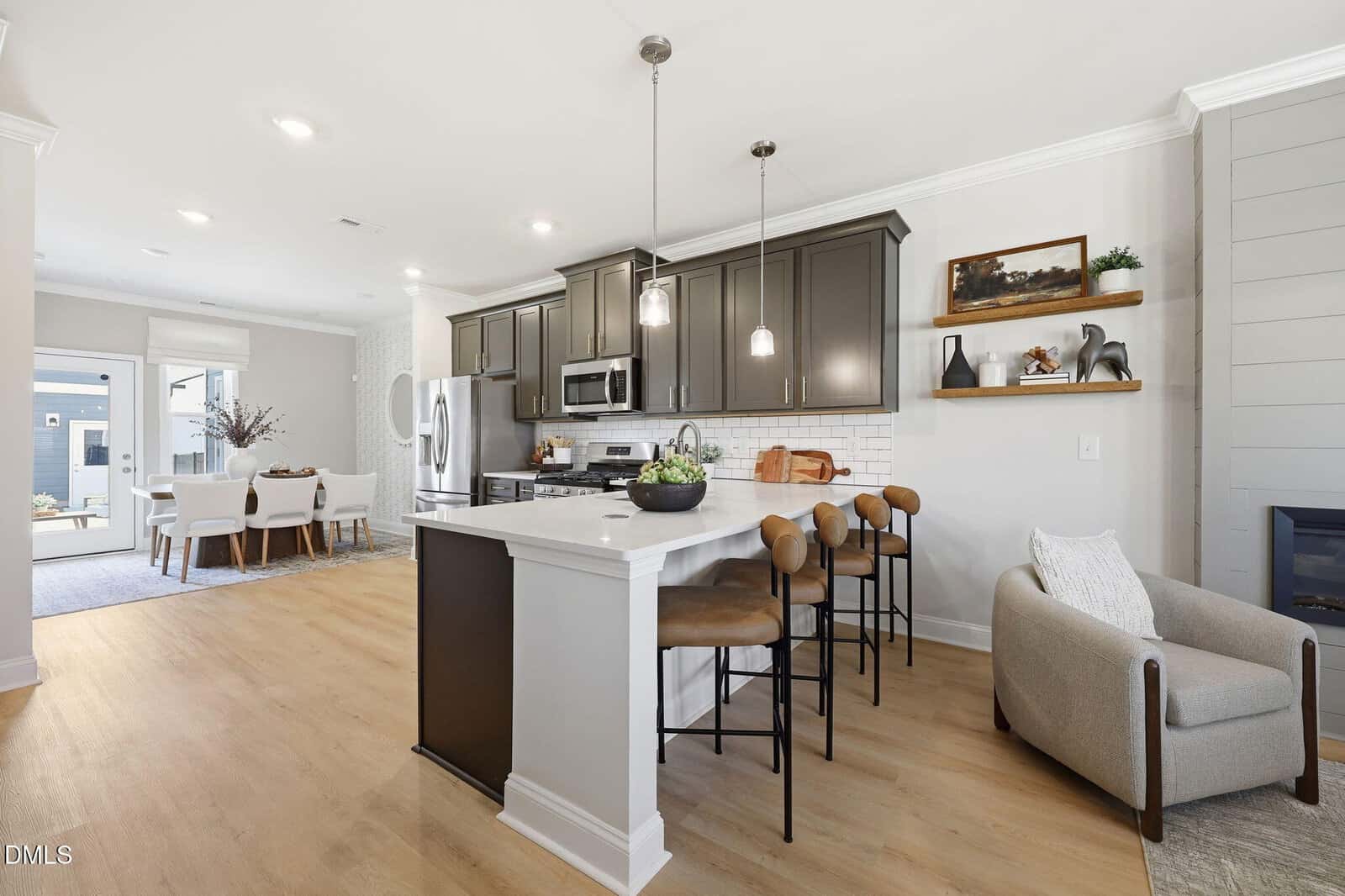 Modern kitchen with gray cabinets, stainless steel appliances, a white island with brown barstools, pendant lights, and open shelving. Adjacent dining area and cozy seating nook with an armchair are also visible.