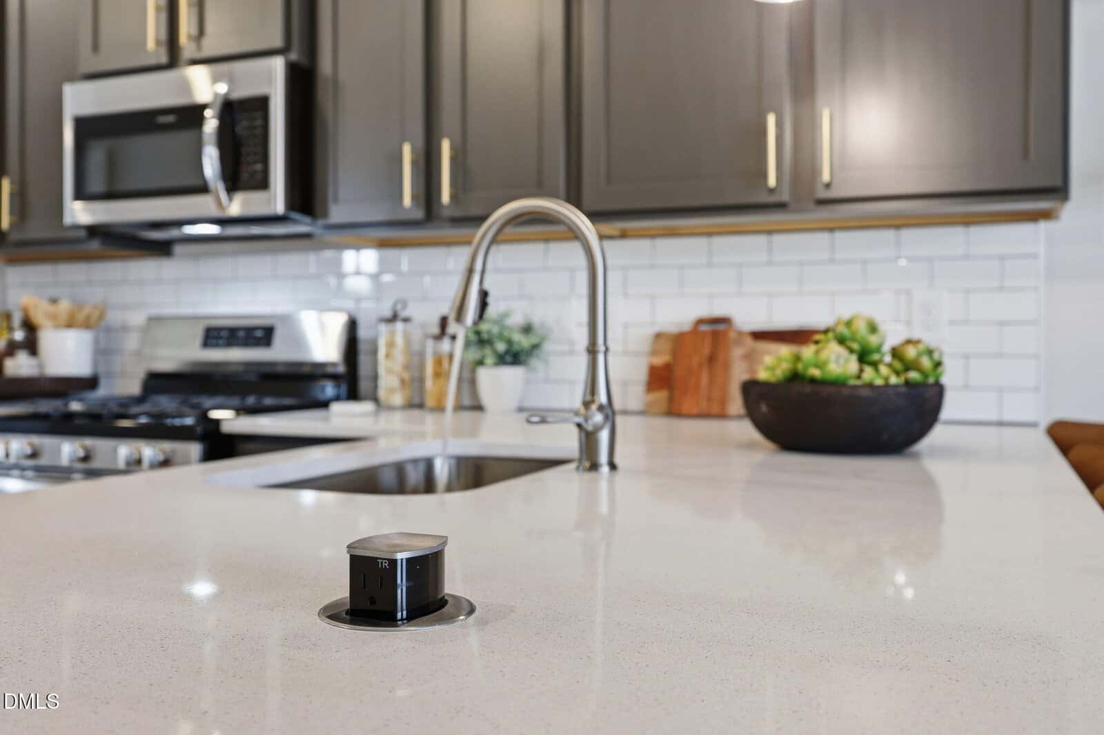 Modern kitchen with gray cabinets, stainless steel appliances, white backsplash, and a light countertop. A pop-up electrical outlet is installed in the counter near a sink with a running faucet. A bowl of artichokes is visible.