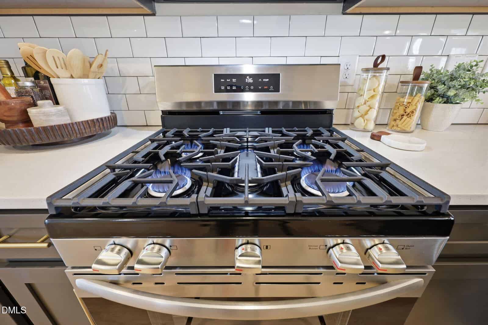 A stainless steel gas stove with four burners, three of which are lit with blue flames. Kitchen utensils, jars, and a spice rack are arranged on the white counter behind the stove.