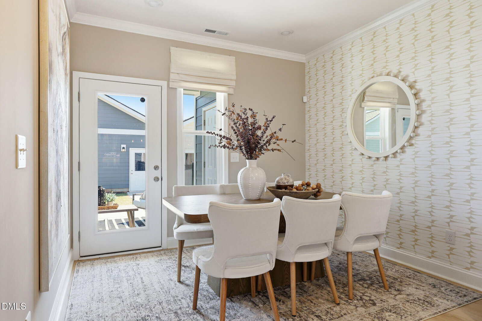 A modern dining room with a wooden table, six white upholstered chairs, a large vase with dried branches, a bowl of decorative objects, a wall mirror, patterned wallpaper, and a glass door leading outside.
