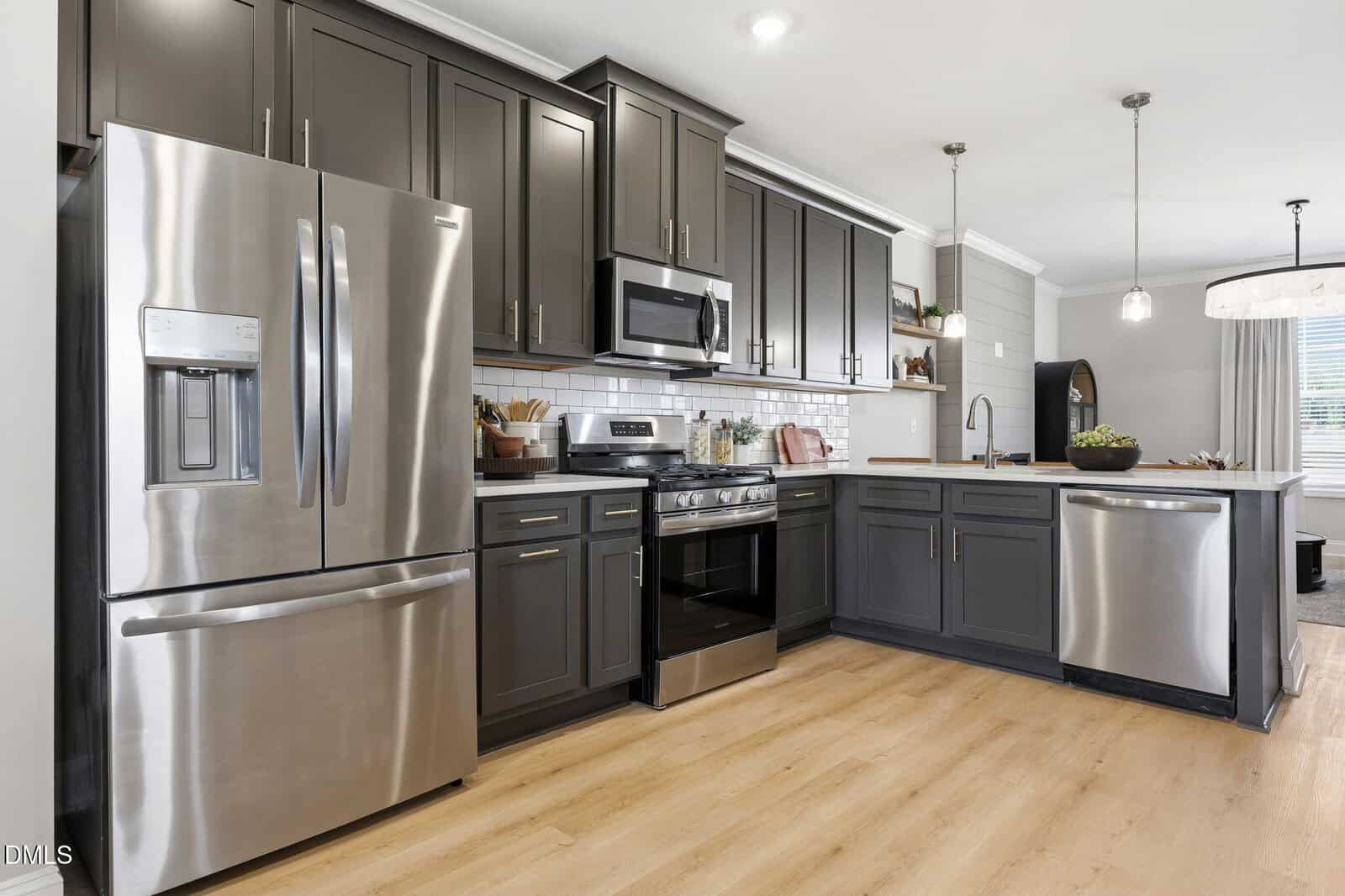 Modern kitchen with dark gray cabinets, stainless steel appliances including refrigerator, oven, microwave, and dishwasher, white subway tile backsplash, light wood floor, and an island with sink.