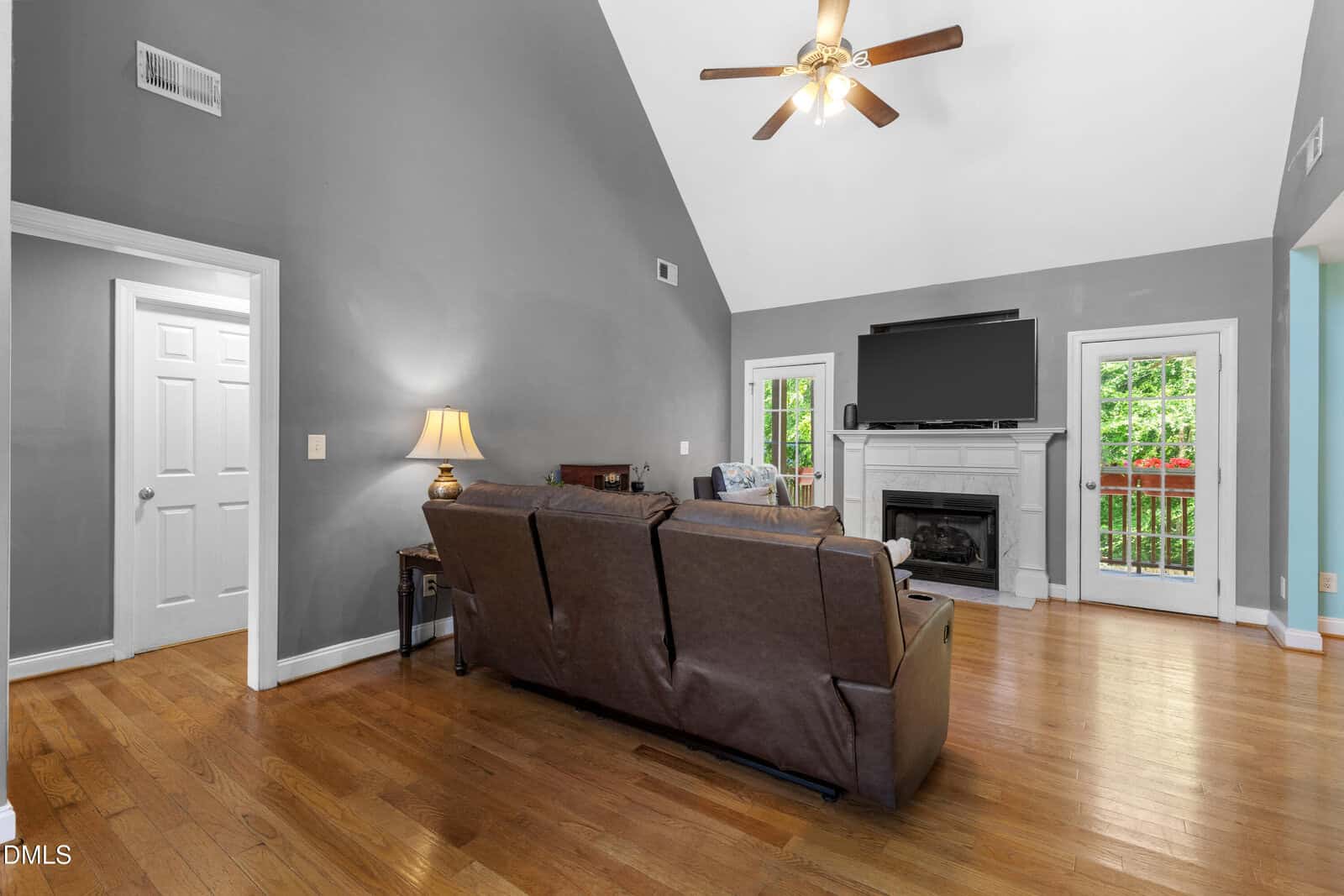 A living room with a brown sofa, hardwood floors, gray walls, a ceiling fan, a fireplace with a TV above, a lamp, and French doors leading to a green outdoor area.