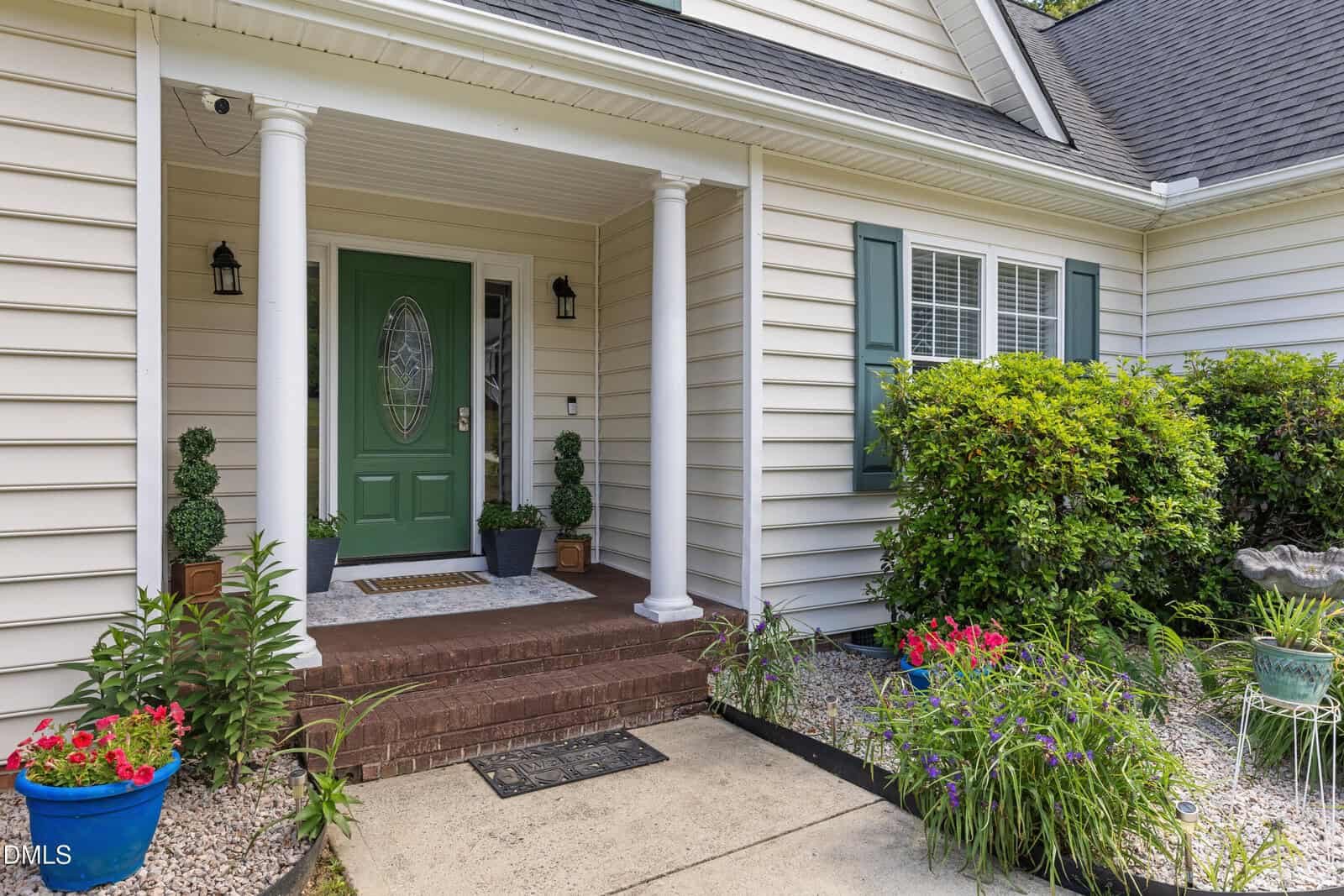 Front porch of a house with a green door, white columns, beige siding, potted plants on the steps, flowers in garden beds, and a window with white shutters to the right.