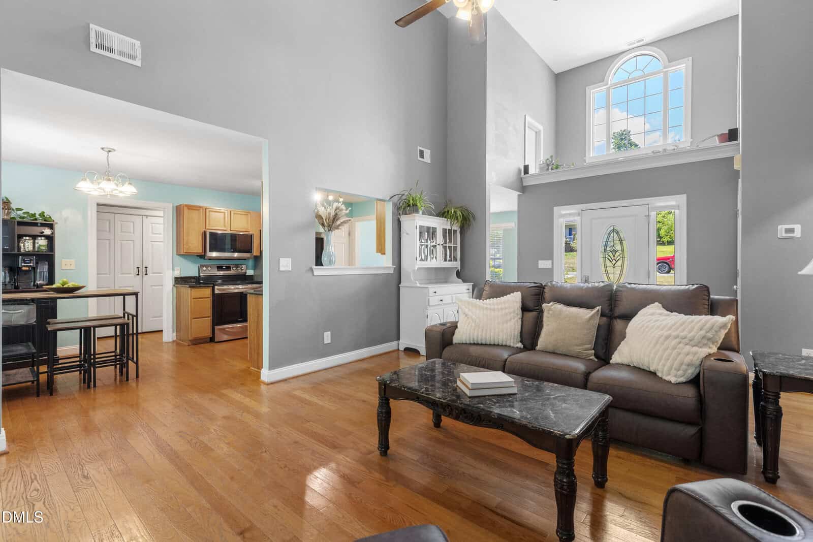 A bright living room with hardwood floors, brown sofa, black coffee table, and high ceiling. Kitchen and dining area are visible. Large window above door lets in natural light.