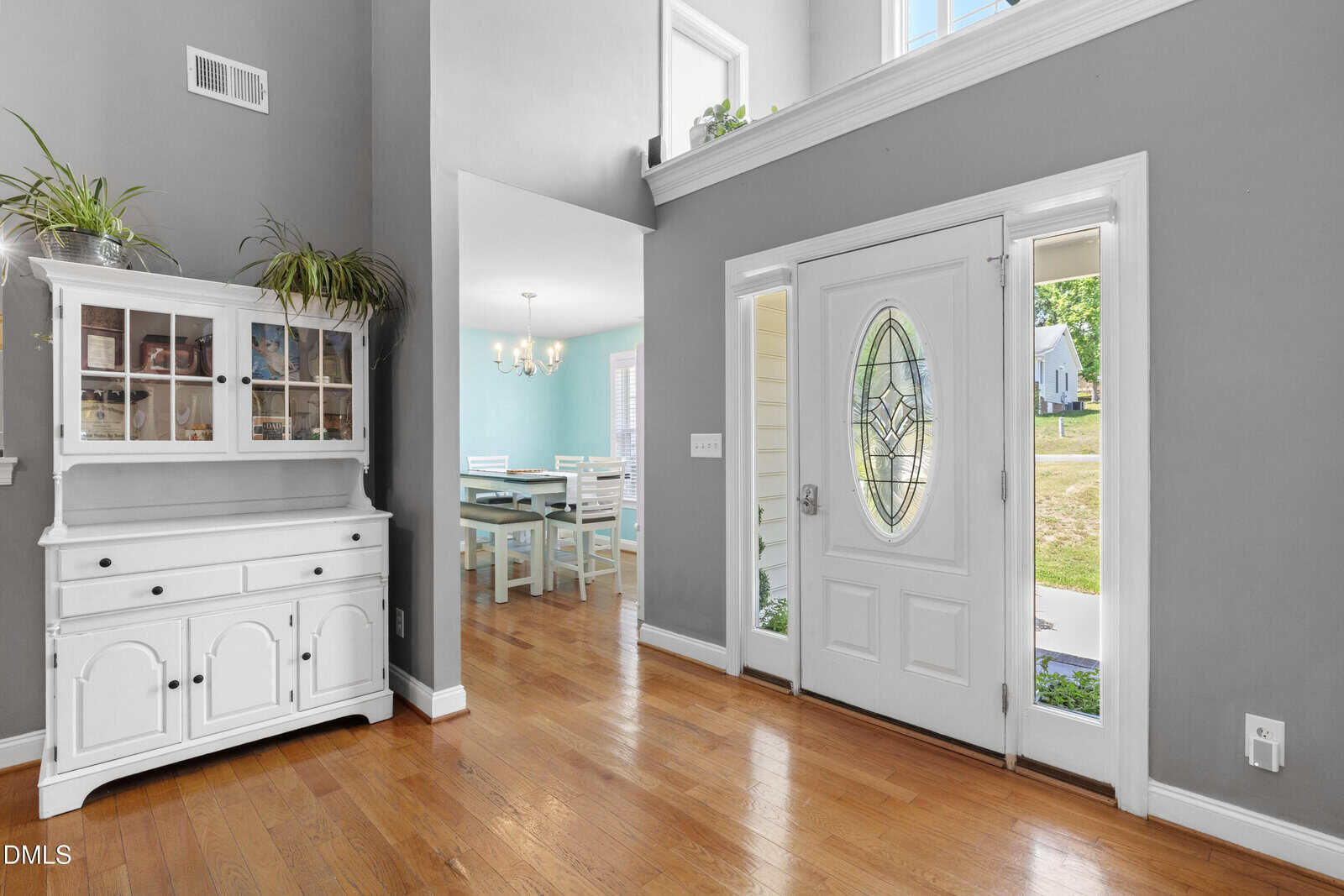 Bright entryway with a white oval glass front door, sidelights, hardwood floors, a white hutch with plants, and a view into a dining room with blue walls and white furniture. Large windows let in natural light.