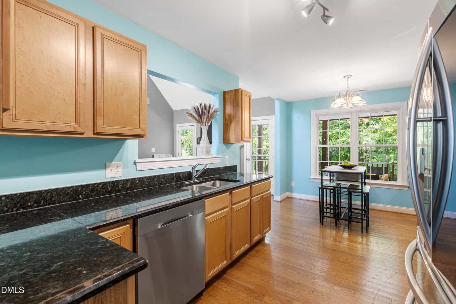 Bright kitchen with light wood cabinets, black granite countertops, stainless steel appliances, and light blue walls. Breakfast nook with table and chairs by large windows overlooking greenery. Hardwood floors throughout.