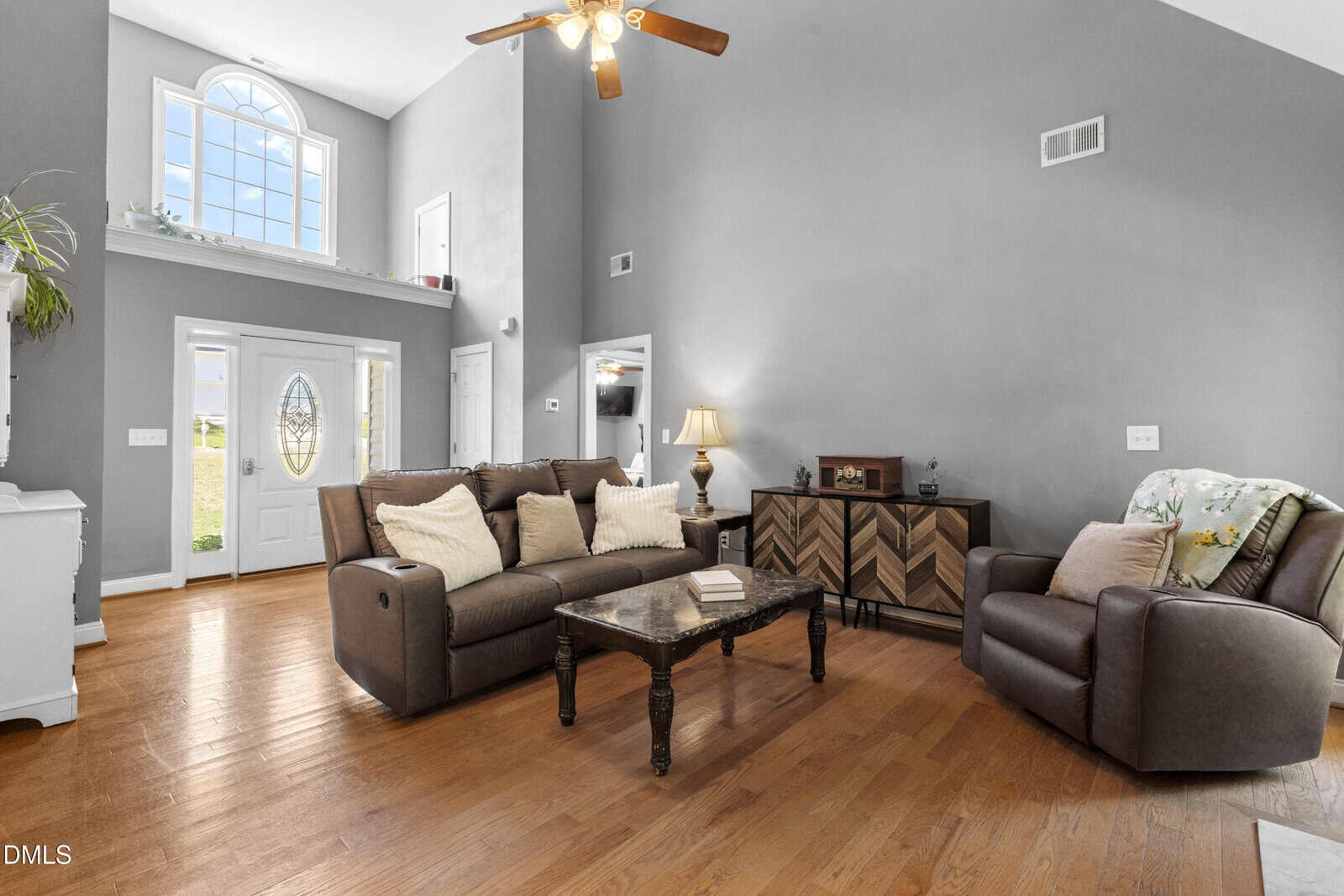 A spacious living room with gray walls, large windows, high ceiling, a ceiling fan, wood floors, a loveseat and armchair with pillows, a coffee table, and a sideboard; front door and staircase visible.