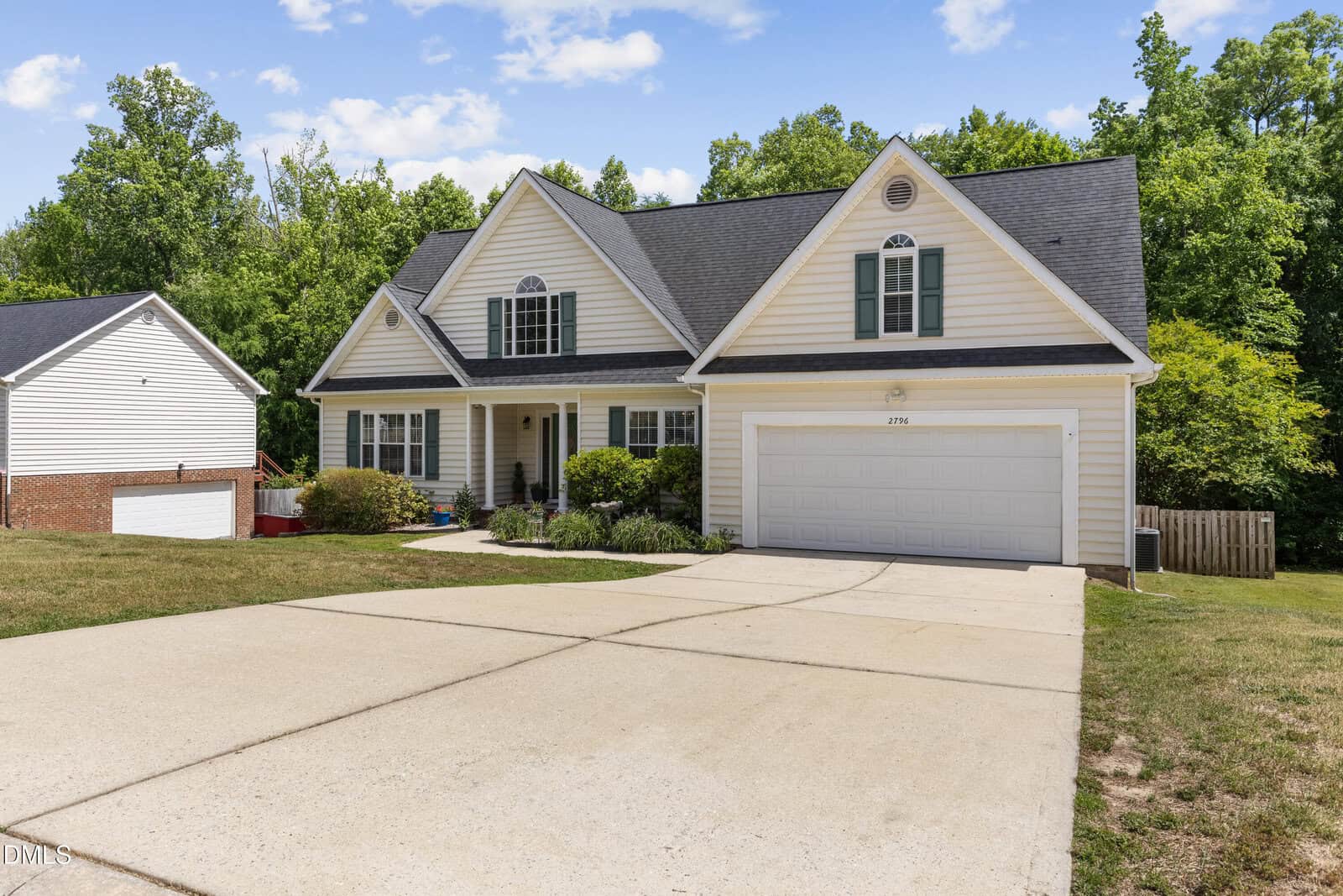 A two-story suburban house with white siding, green shutters, a large driveway, and an attached double garage, surrounded by trees and a well-kept lawn under a blue sky with clouds.