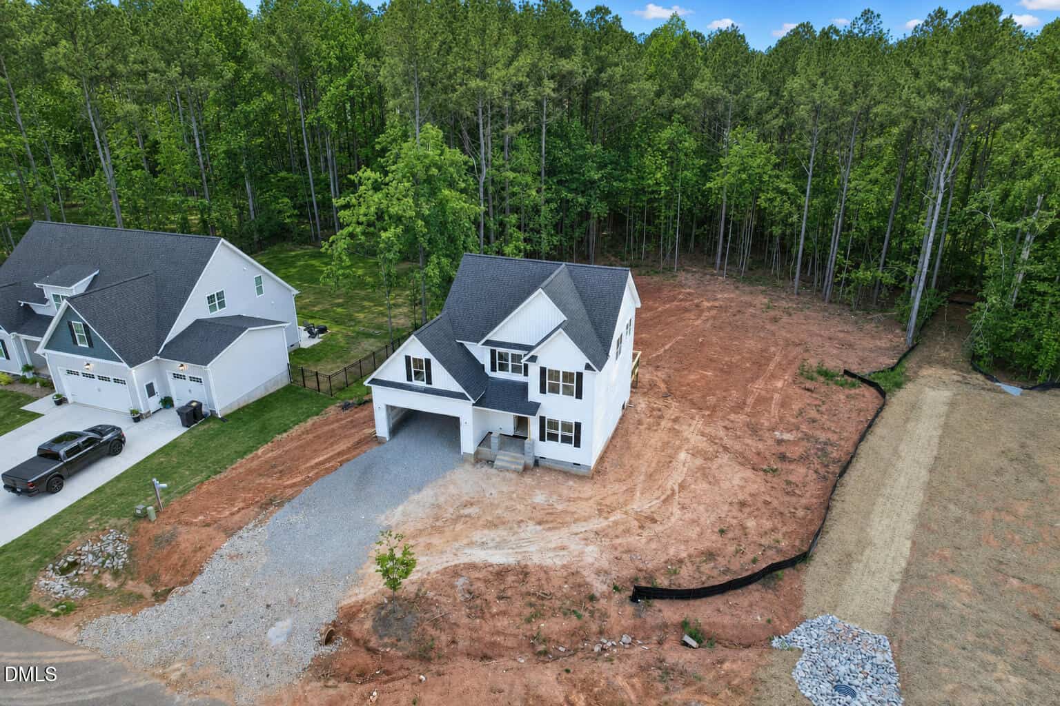 A newly built two-story white house with a gray roof sits on a large dirt lot next to a similar house, surrounded by trees. There is a driveway, minimal landscaping, and construction materials on site.