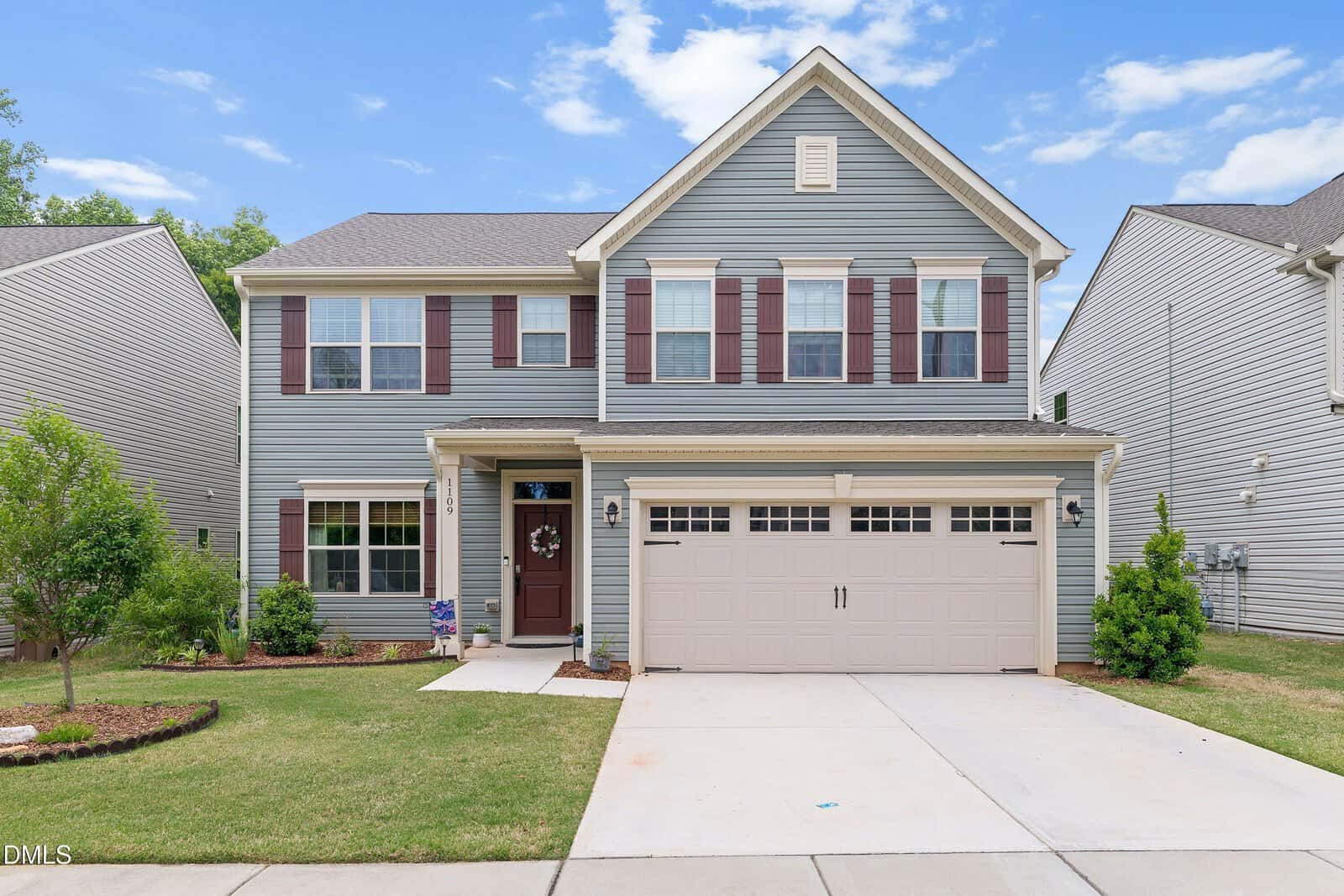 Charming two-story suburban house at 1109 Spring Meadow Way in Wake Forest, featuring blue-gray siding, white trim, maroon shutters, a double garage, manicured lawn, and inviting landscaping under a blue sky with clouds.