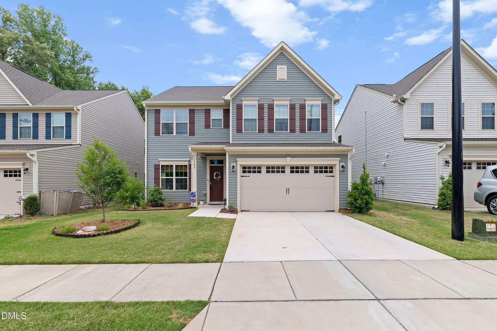 Two-story blue house with white trim, red shutters, and a double garage. Well-maintained lawn and landscaping; neighboring houses on both sides; clear daytime sky with scattered clouds.