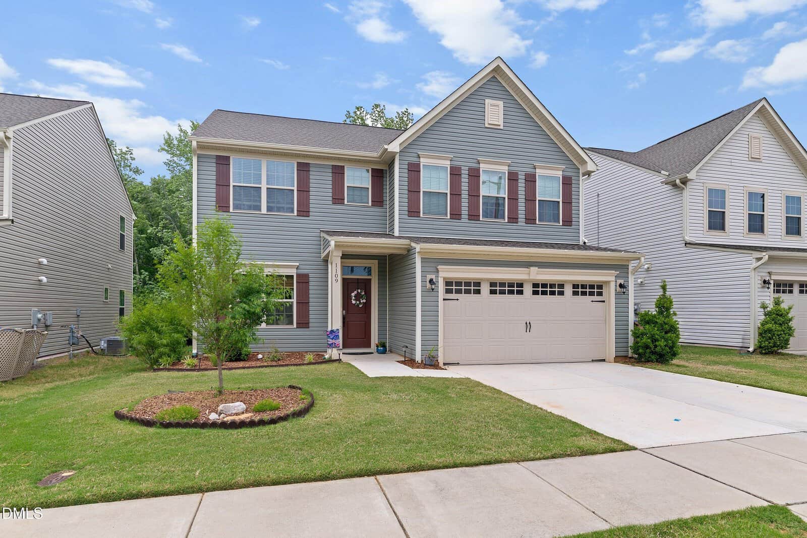 Two-story gray house with white trim and maroon shutters, attached two-car garage, small covered front porch with a wreath, and a well-kept lawn with minimal landscaping. Sky is clear with a few clouds.