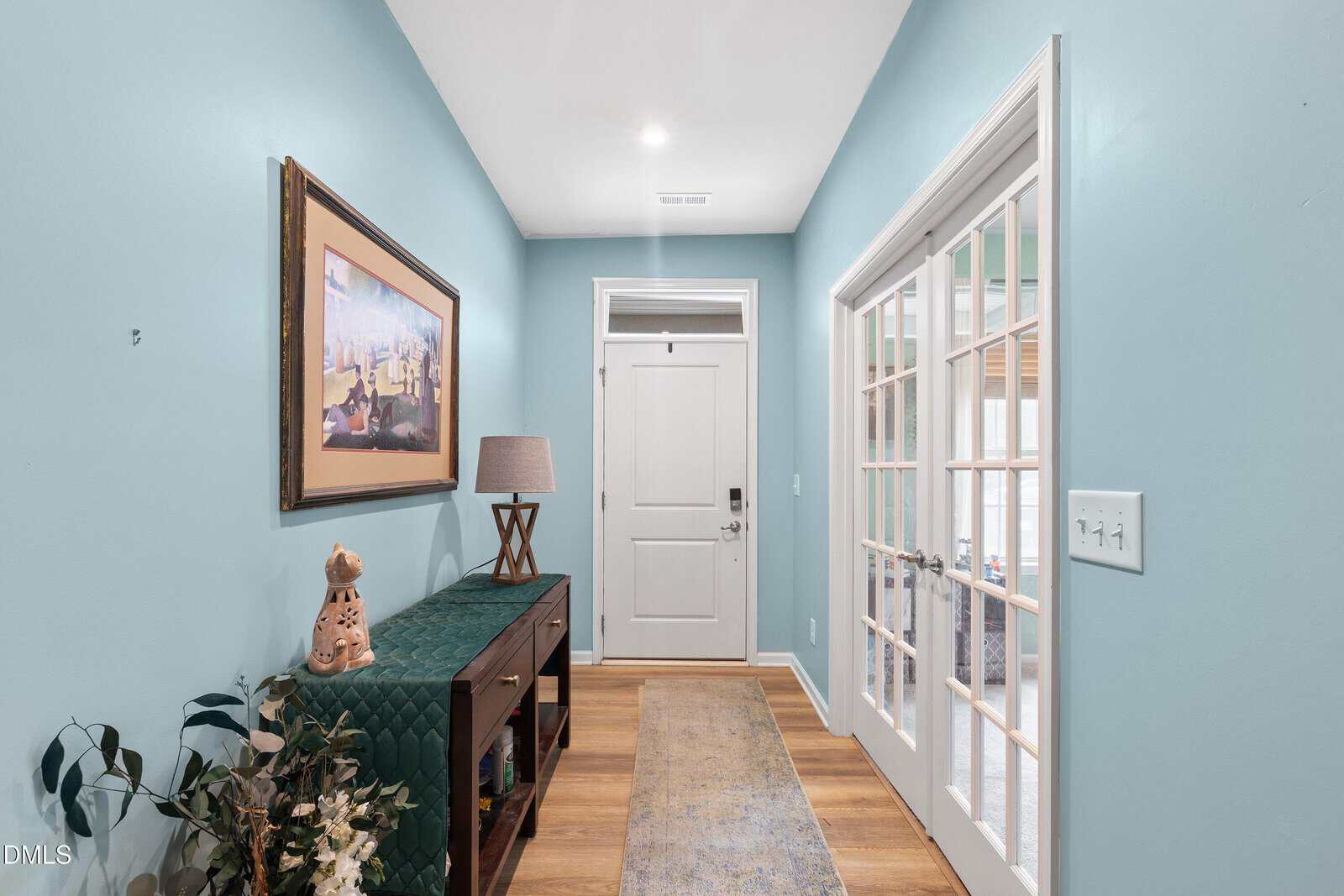 A bright hallway with light blue walls, wood flooring, a white front door, a rug, a glass-paned double door, a console table with a lamp, decorative items, and a framed painting on the wall.