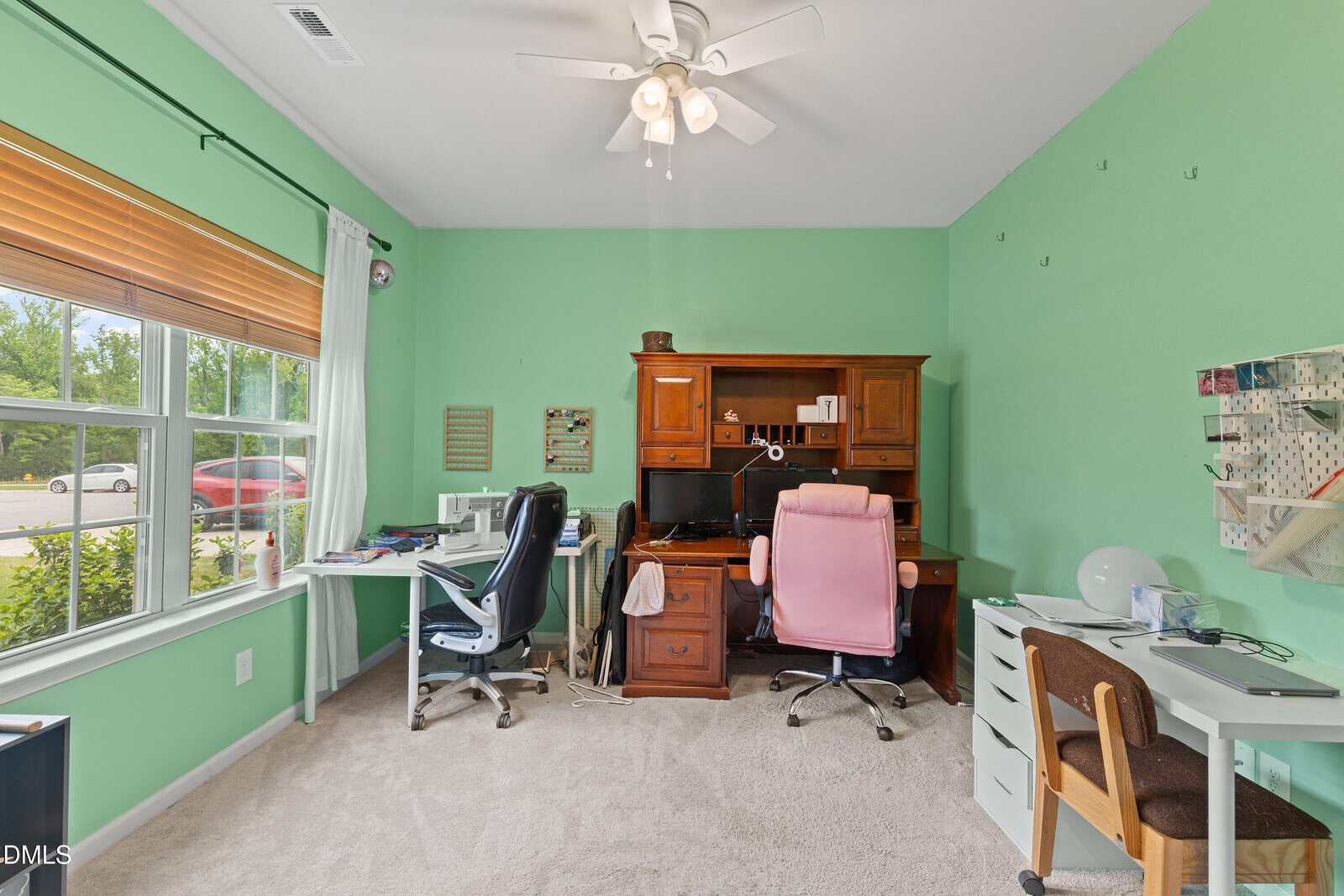 Home office with mint green walls, three desks with chairs, a wooden computer hutch, a sewing machine, and various office supplies. Two windows with blinds let in natural light. Carpeted floor and ceiling fan are visible.