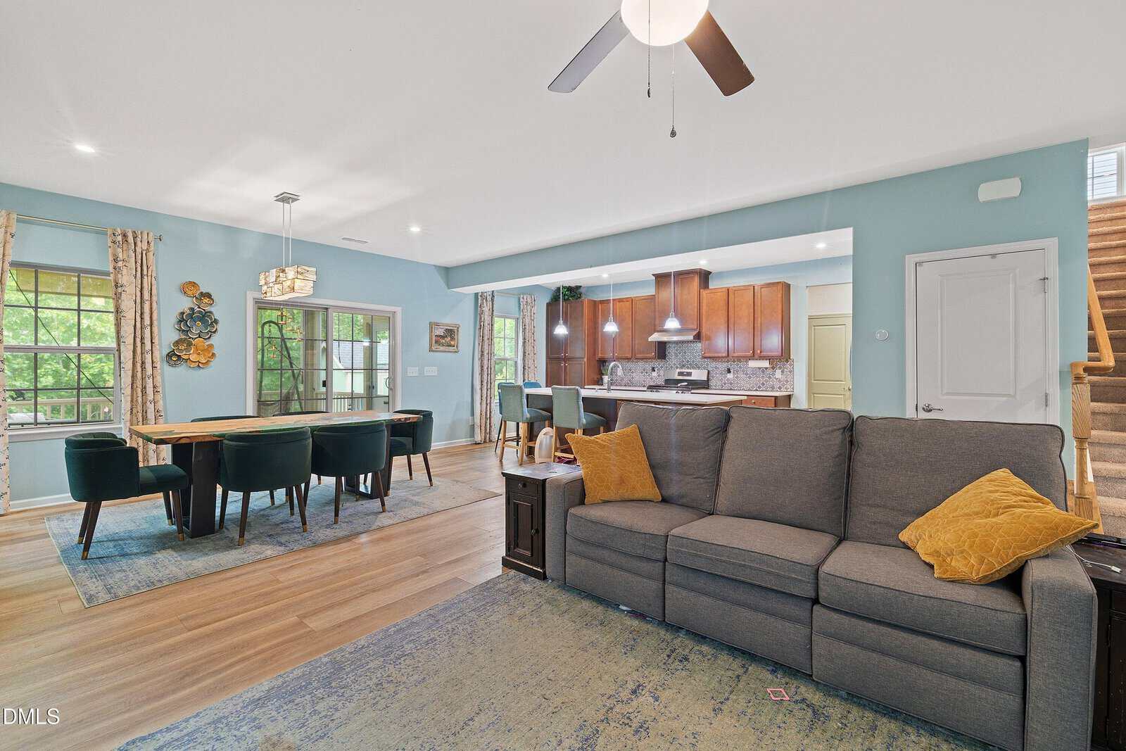 Open living room and dining area with light blue walls, wooden floors, modern gray sofa, green dining chairs, and a kitchen with brown cabinets in the background. Stairs are visible on the right.