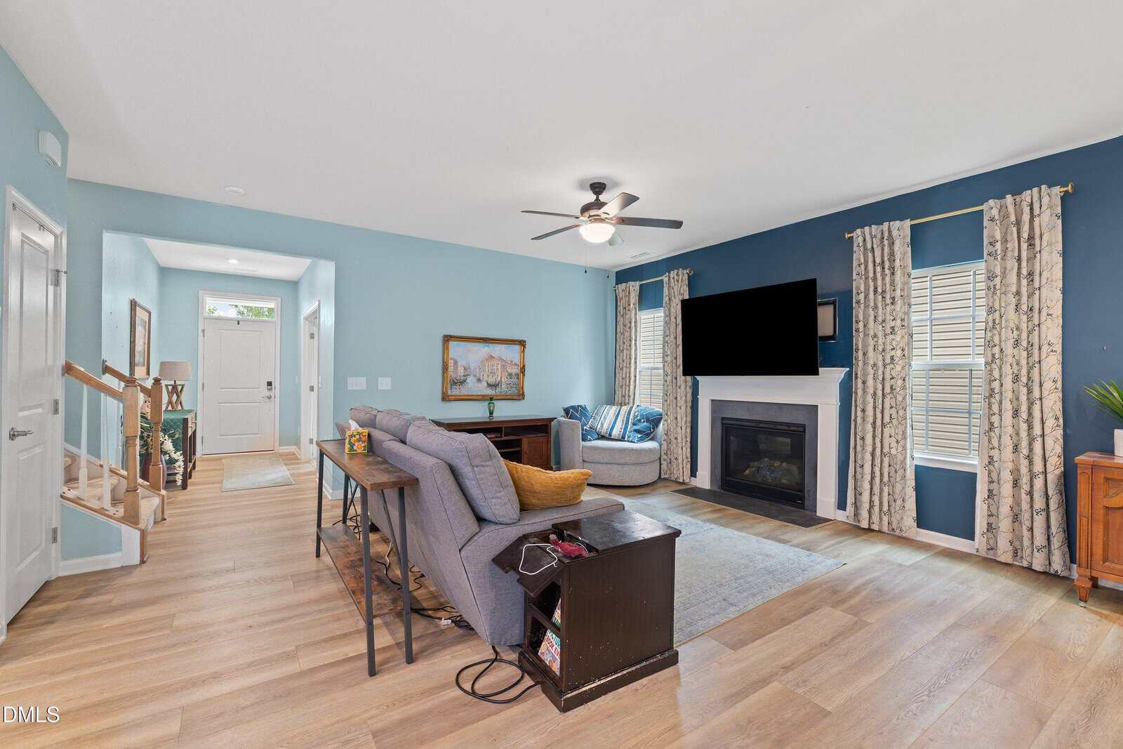 A cozy living room with blue walls, a gray sectional, TV above a white fireplace, wood floors, windows with patterned curtains, and an entryway leading to a staircase.