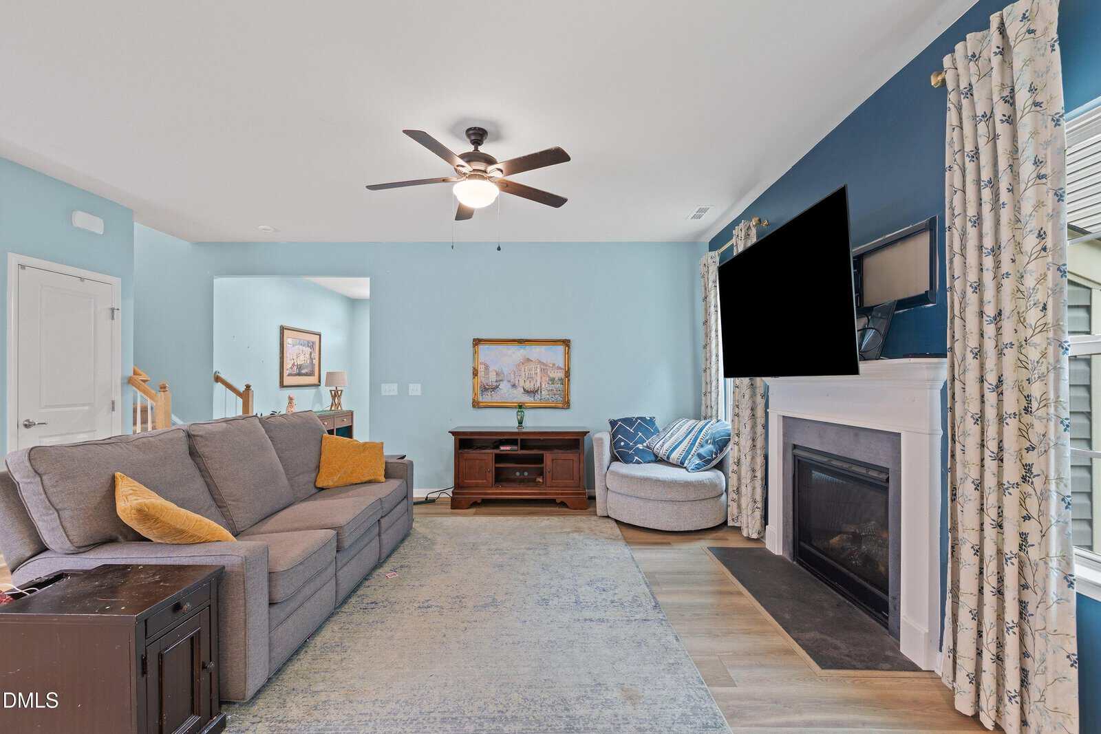 A cozy living room with a gray sectional sofa, wall-mounted TV above a fireplace, blue accent wall, round chair, and large windows with patterned curtains. Ceiling fan and side table are also visible.