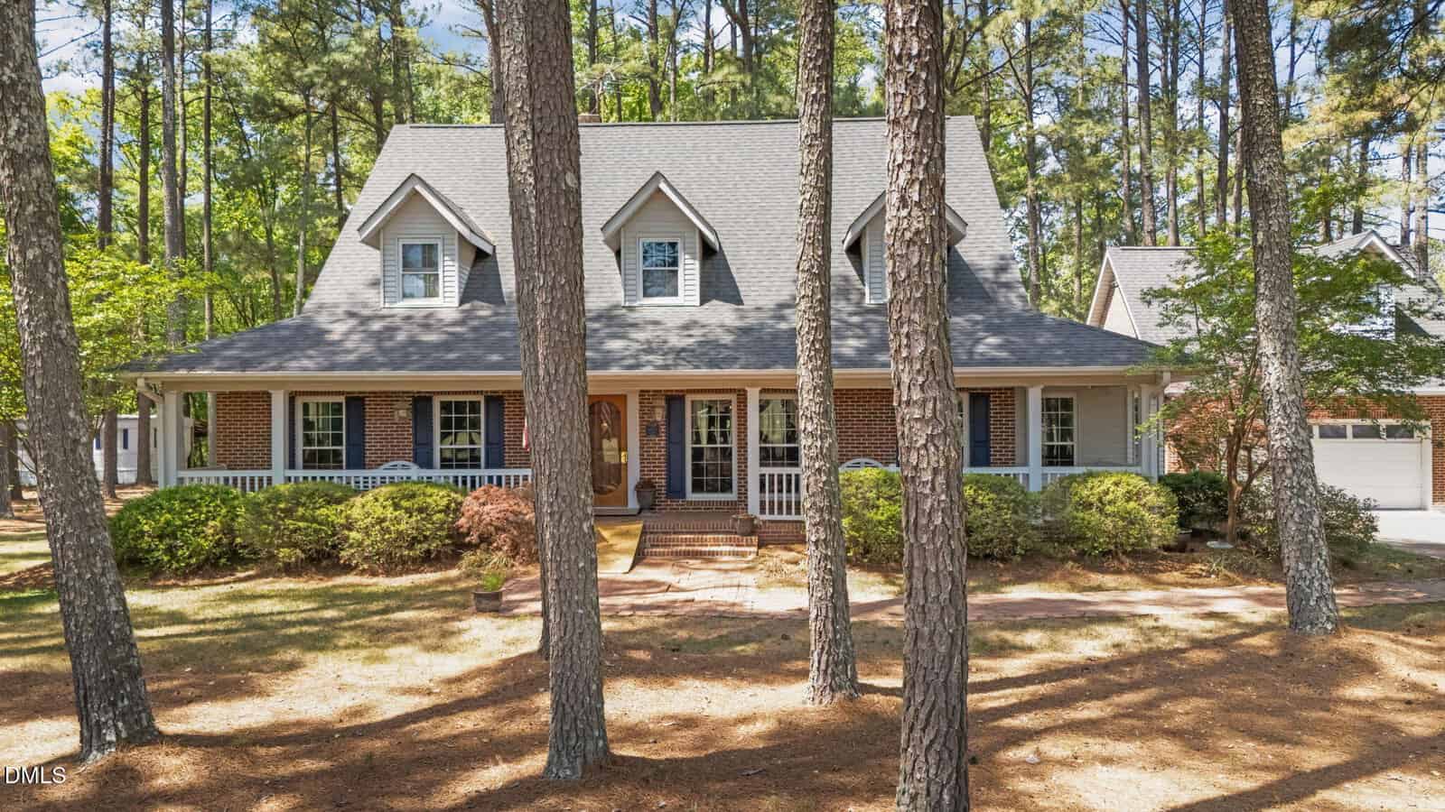 A two-story brick house in Oxford at 4509 S Highway features a large front porch, dormer windows, and a gabled roof, nestled among tall pine trees and landscaped shrubs in the front yard.