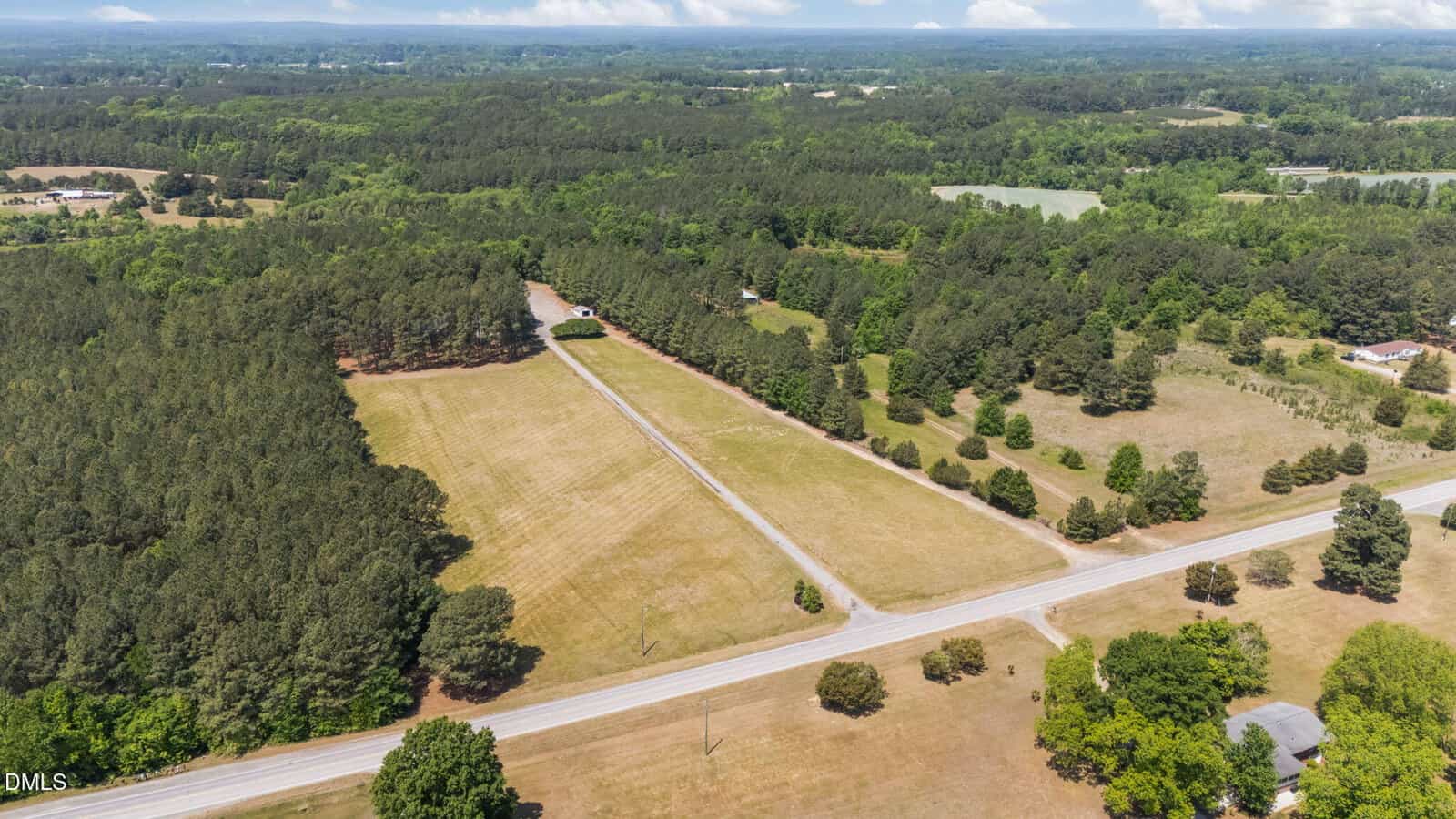 Aerial view of a rural landscape with a long driveway leading to a house surrounded by trees, open fields, and intersecting roads under a partly cloudy sky.