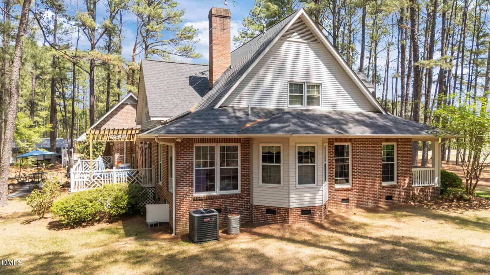 A two-story brick and siding house with a gabled roof, large windows, a bay window, and two HVAC units outside, surrounded by tall pine trees and a shaded lawn.