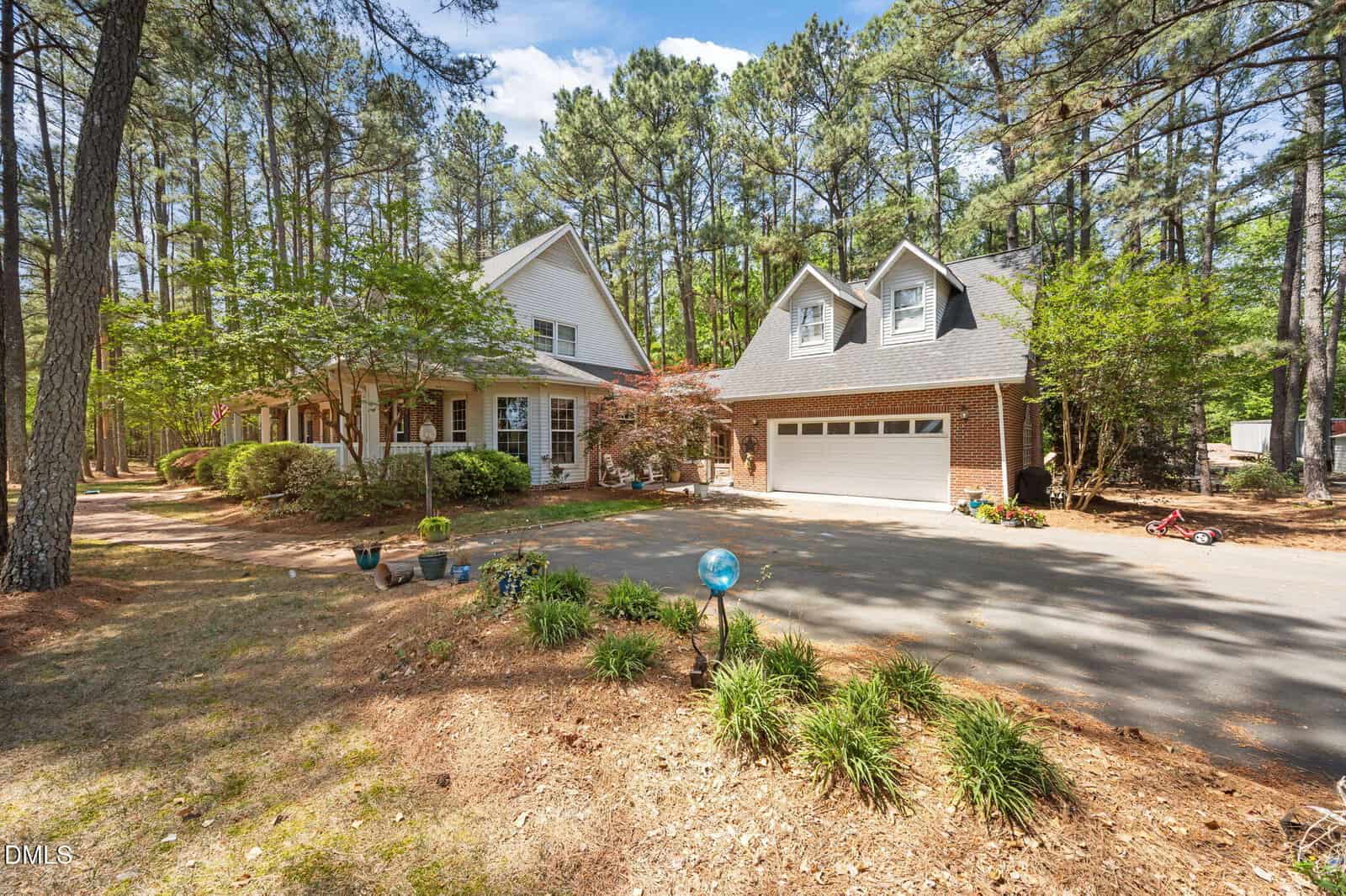A two-story house with a brick garage and white siding sits among tall pine trees, featuring a front porch with columns, a wide driveway, and landscaped garden beds in a wooded setting.