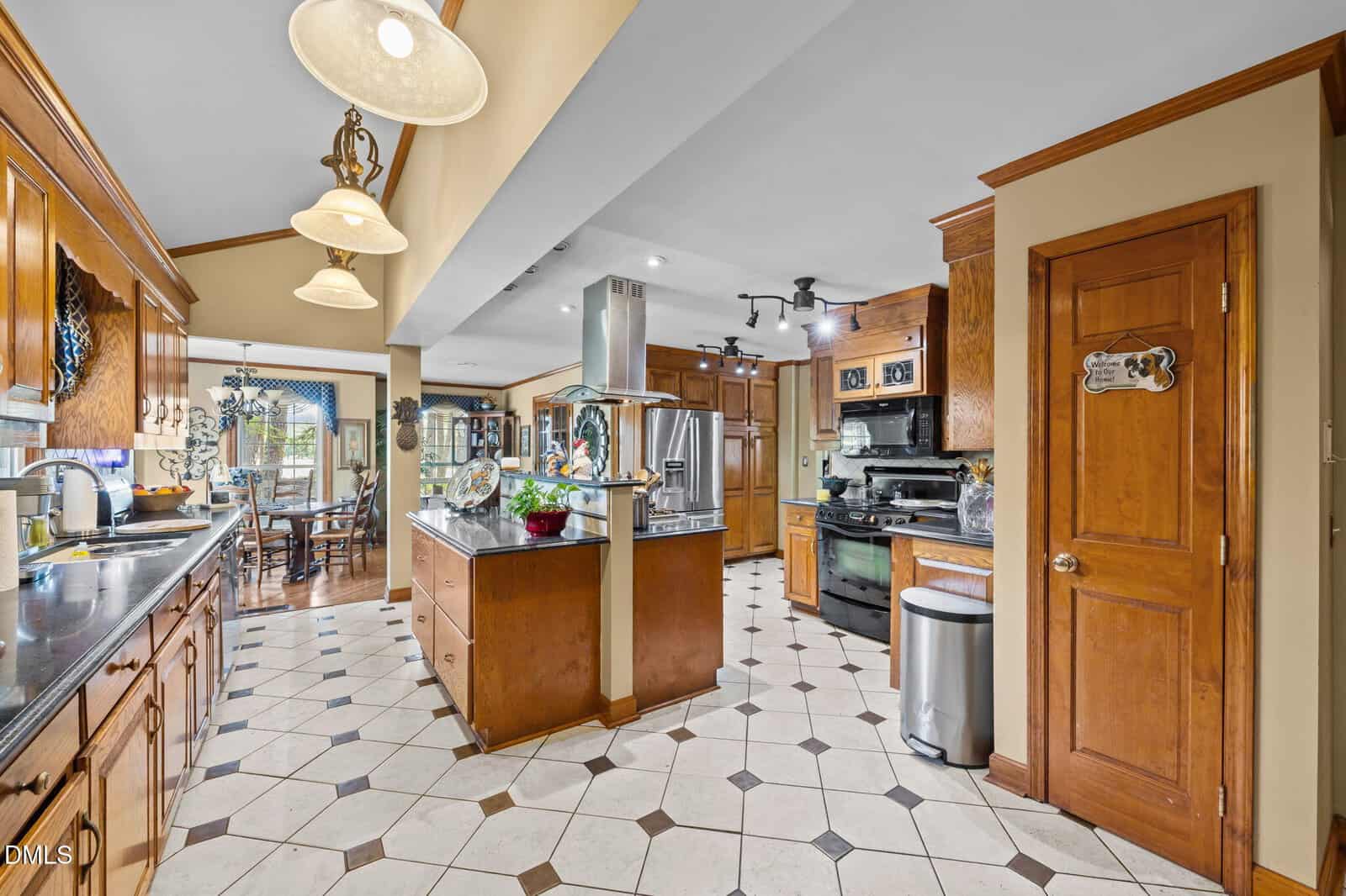 Spacious kitchen with wooden cabinets, stainless steel appliances, a central island, tiled floor, and pendant lighting. Dining area is visible in the background through a wide doorway.
